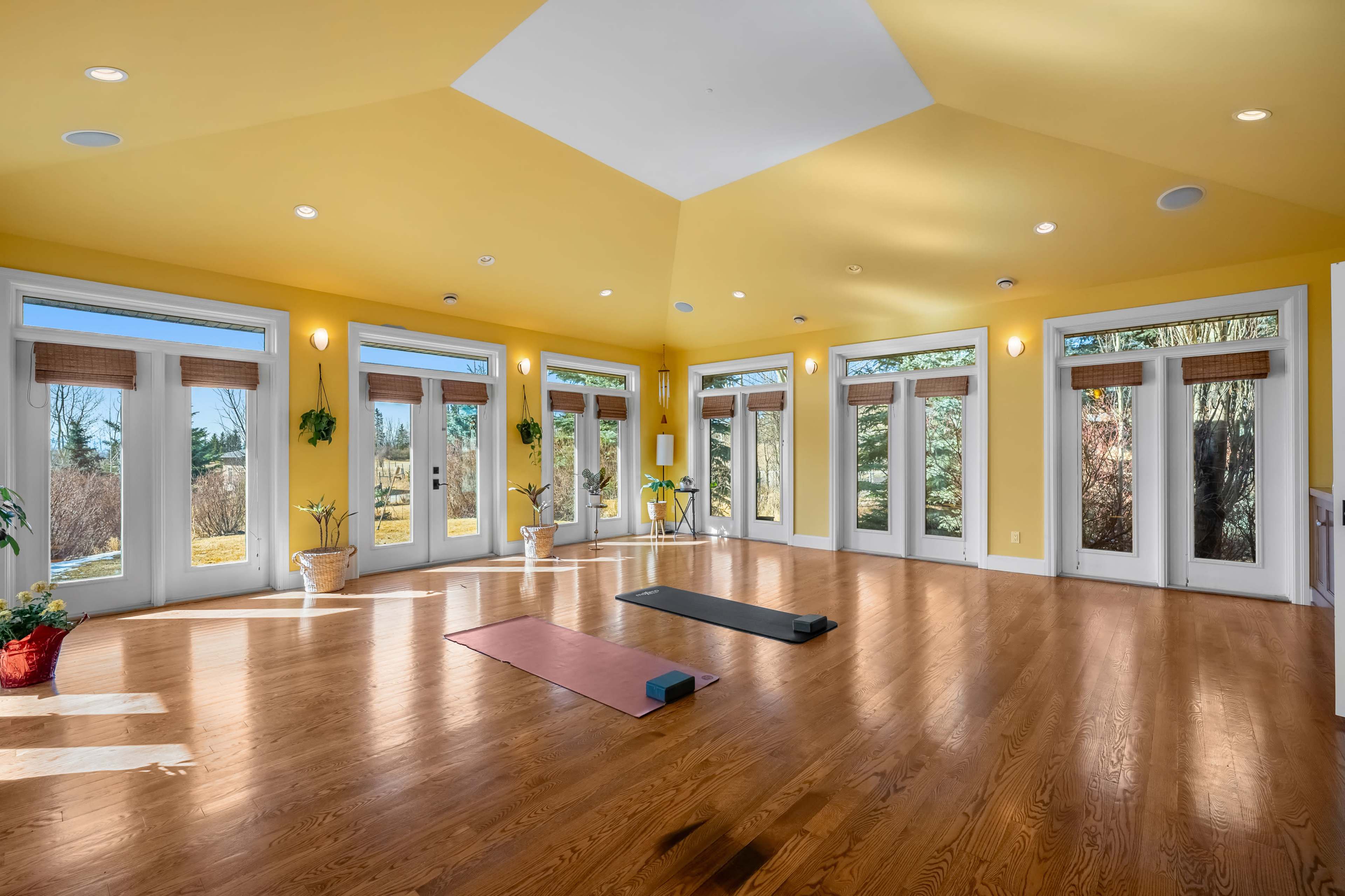 The image shows a bright, spacious yoga studio with large windows allowing natural light to illuminate the wooden floor and two yoga mats on the ground.