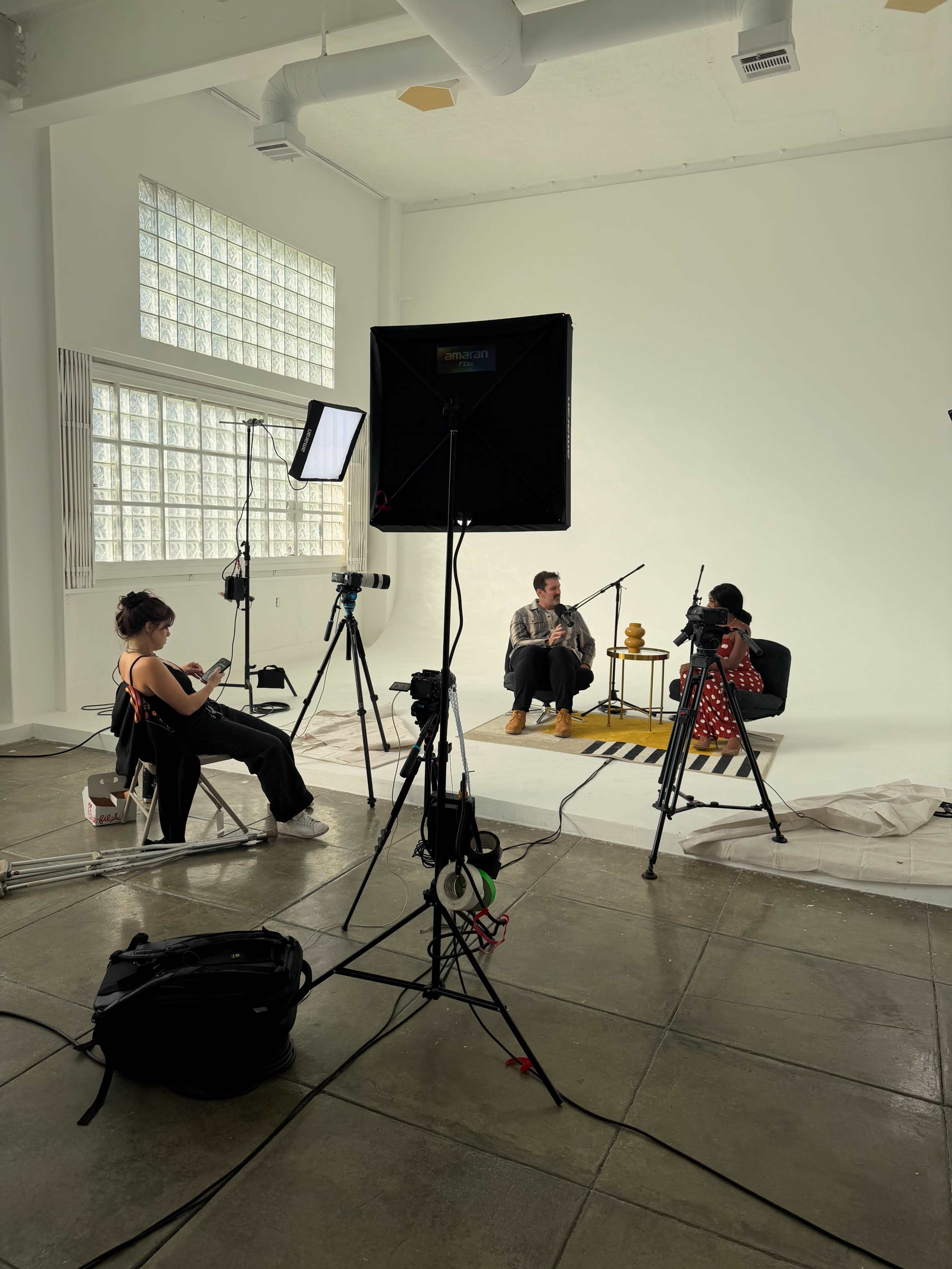 A spacious studio features two people engaged in conversation, surrounded by lighting equipment and cameras set up on tripods.