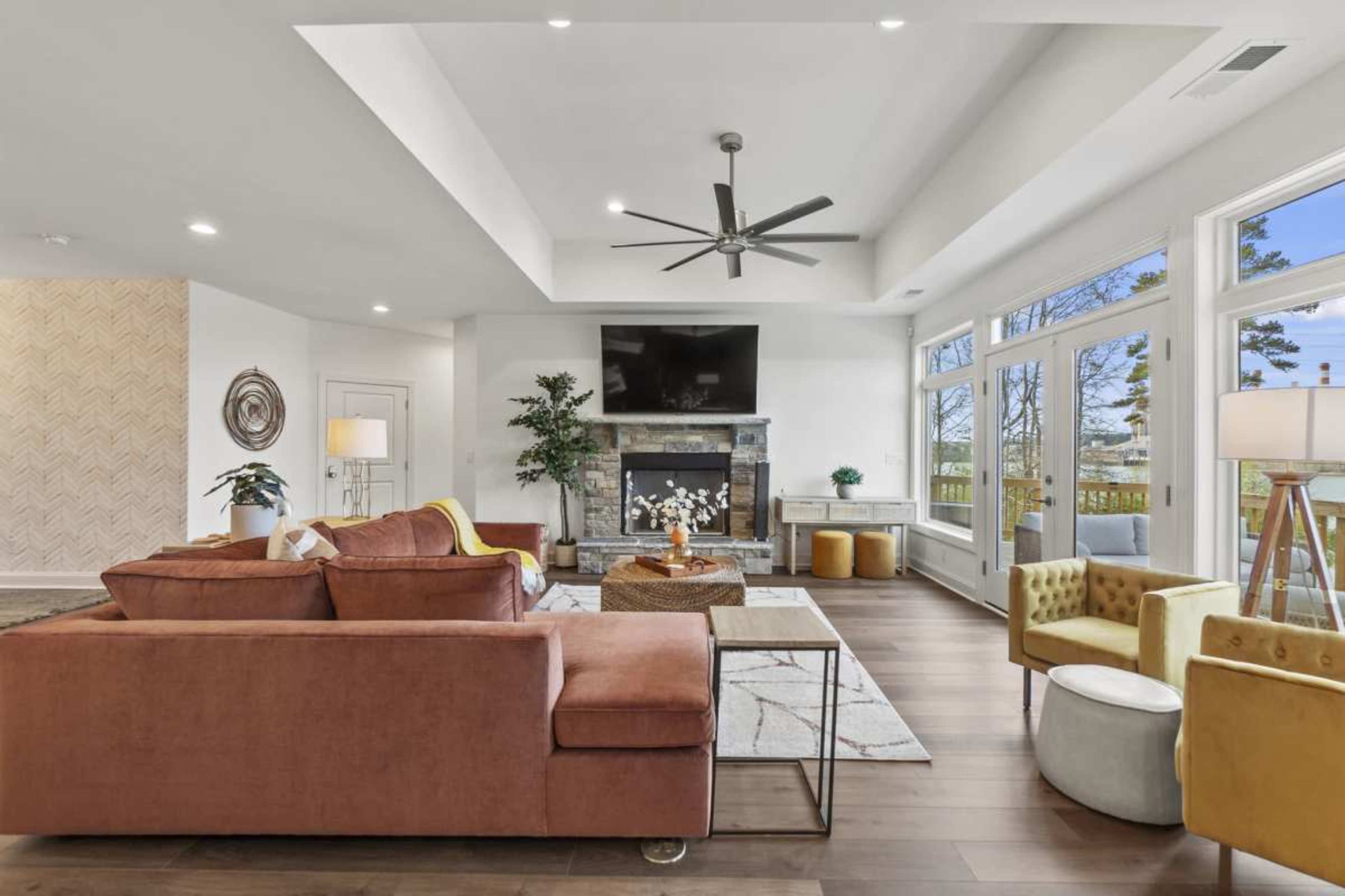 The image shows a modern living room with a large sectional sofa, a stone fireplace, and a television mounted above it, surrounded by large windows that let in natural light.