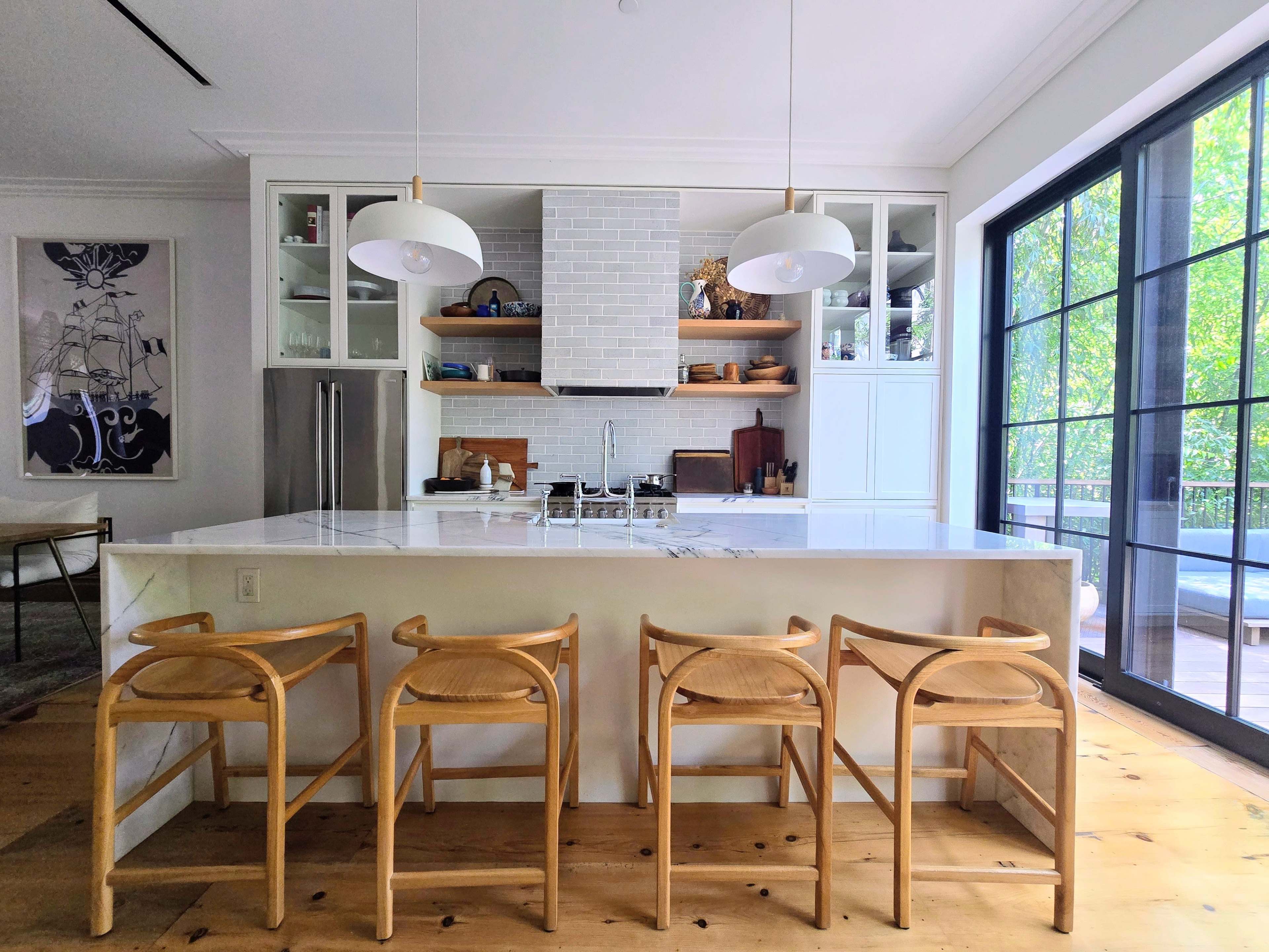 The image depicts a modern kitchen with a large island featuring four wooden stools, a white backsplash, and large windows overlooking a green outdoor space.