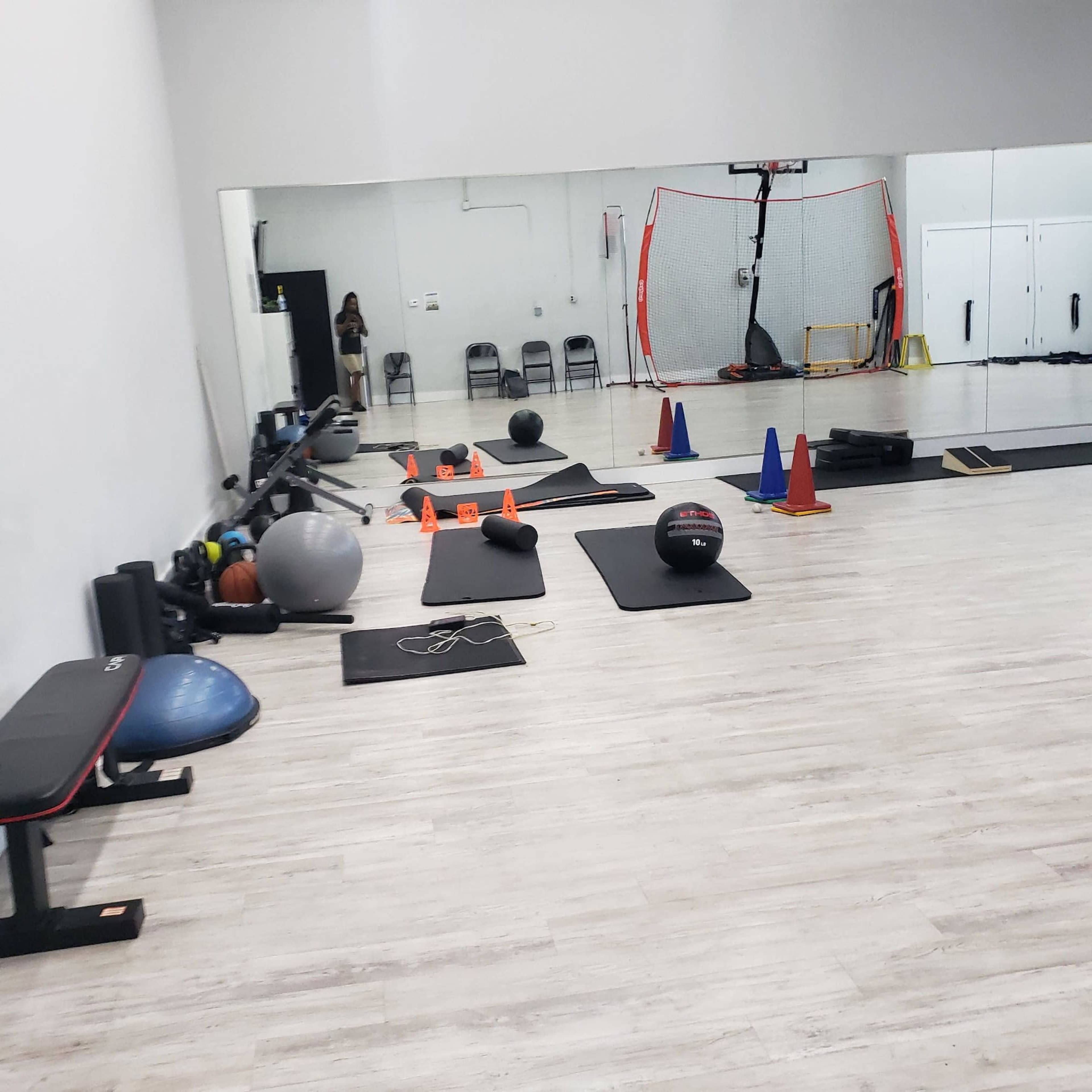 A gym interior with exercise equipment, including mats, exercise balls, cones, and a basketball hoop visible in the background.