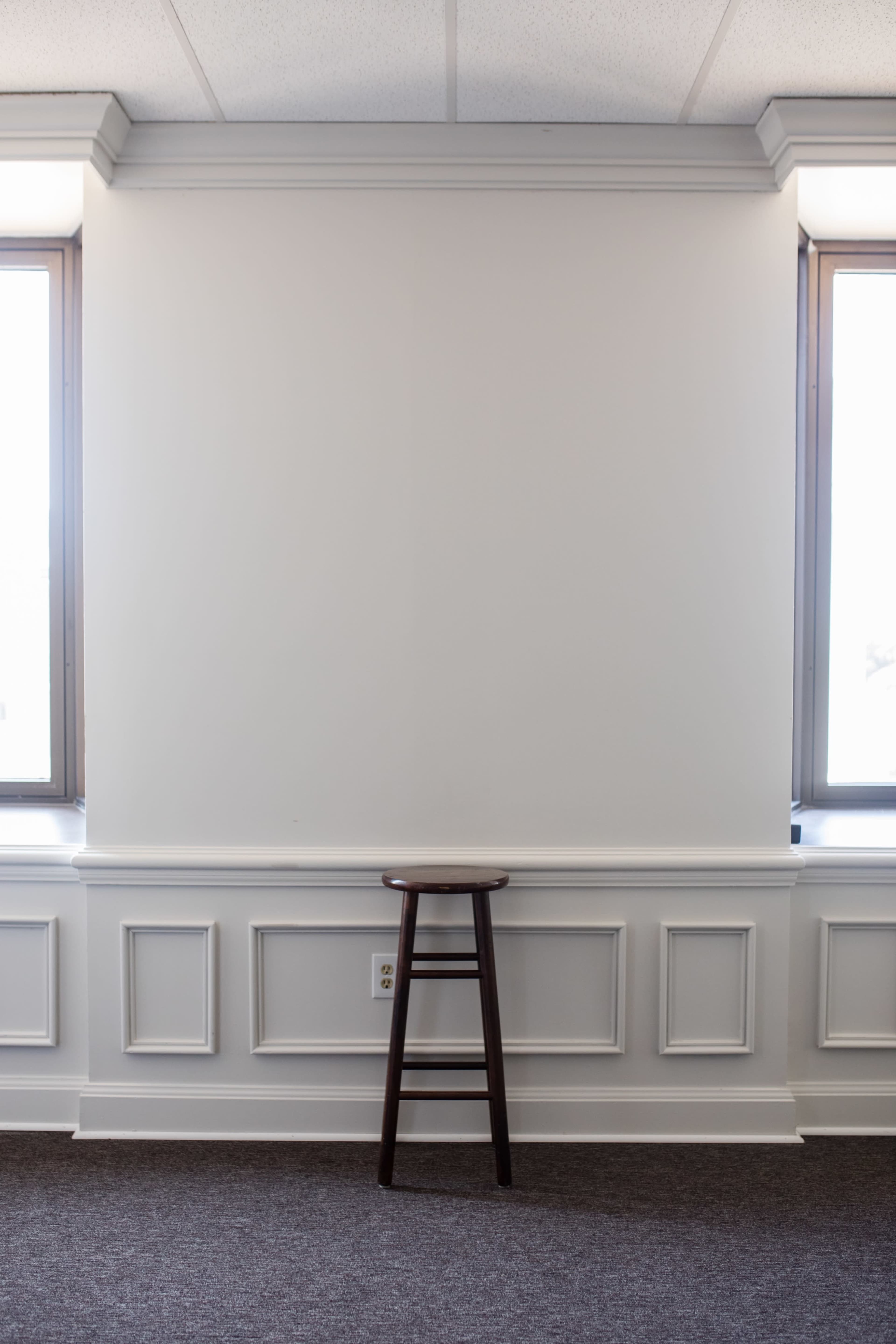 A wooden stool stands against a blank wall between two windows in a room with wainscoting and carpeted flooring.