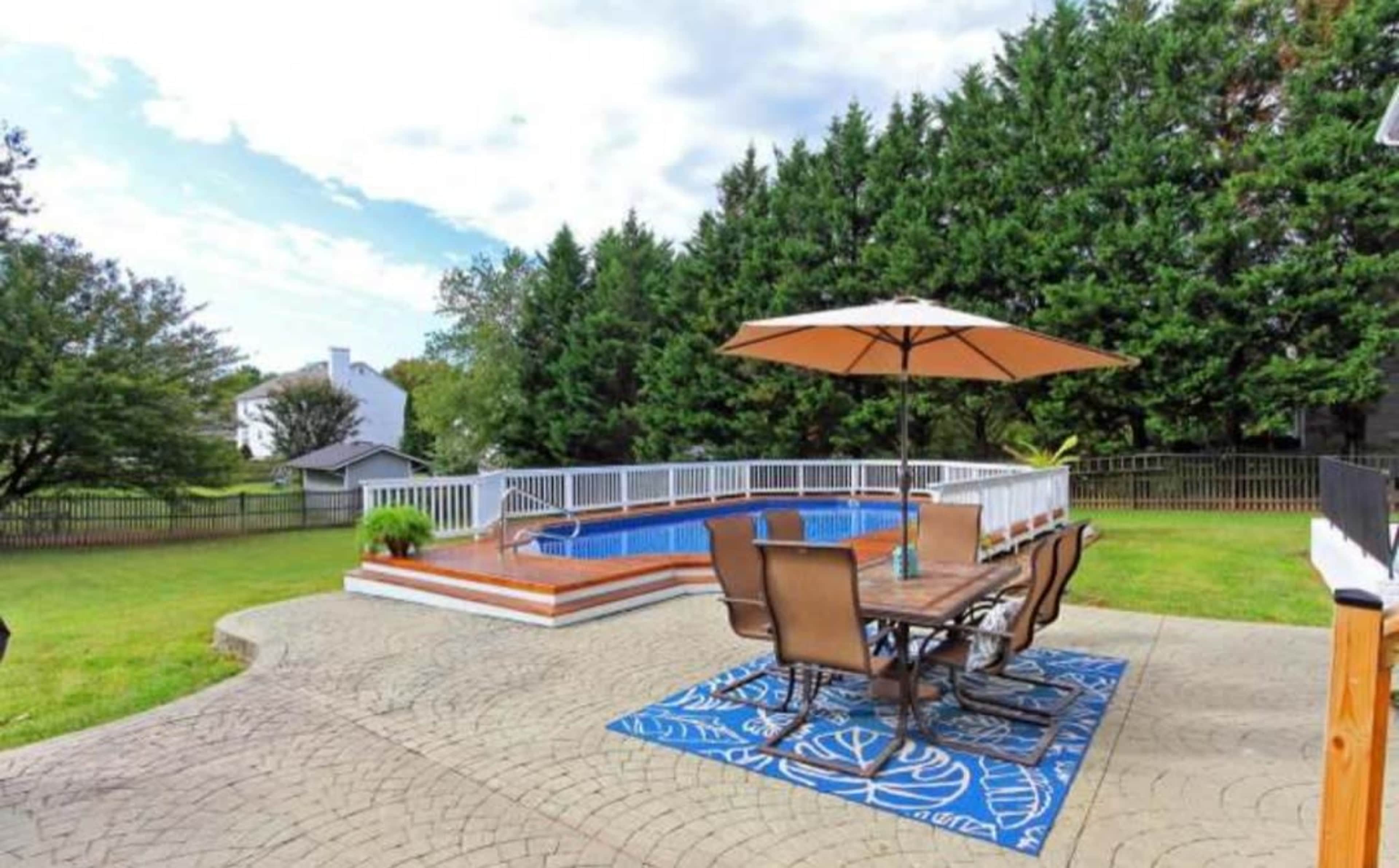 A backyard with a swimming pool surrounded by a patio, outdoor dining furniture, and a large umbrella, bordered by tall greenery.