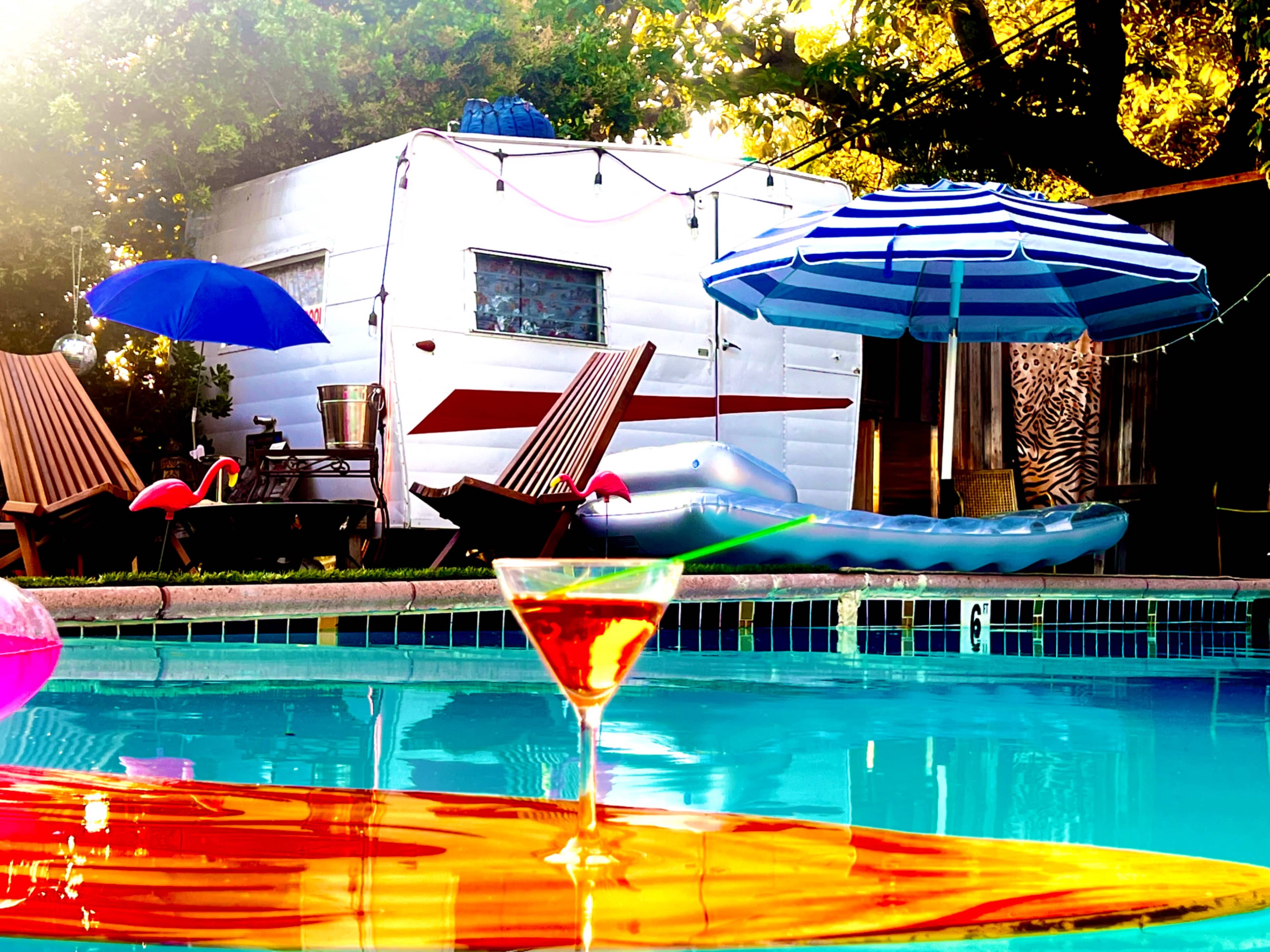 The scene features a colorful swimming pool surrounded by lounge chairs, umbrellas, and a vintage trailer in the background.
