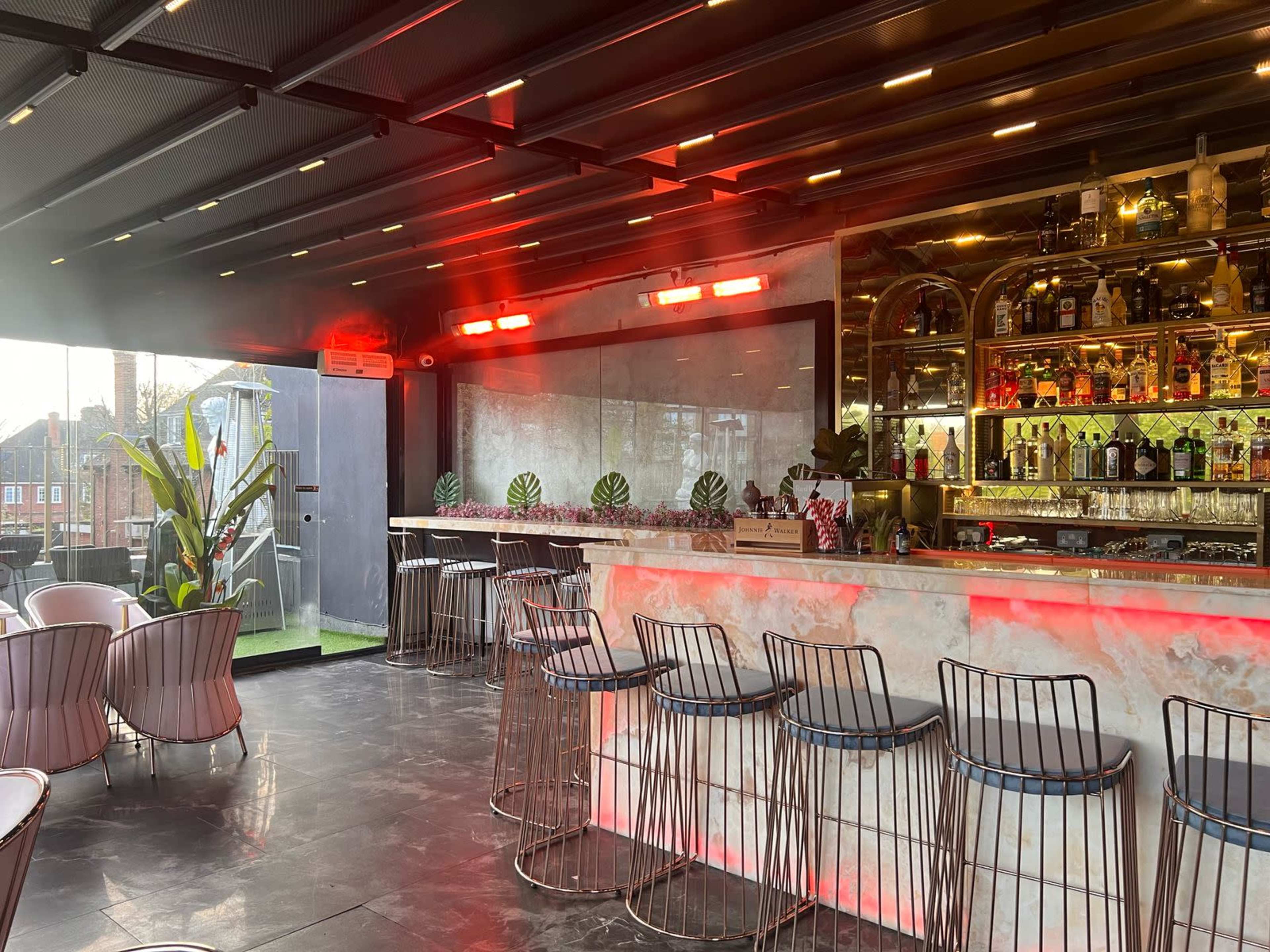 The image shows a modern bar with sleek metal stools, vibrant lighting, and a well-stocked shelf of various liquor bottles.