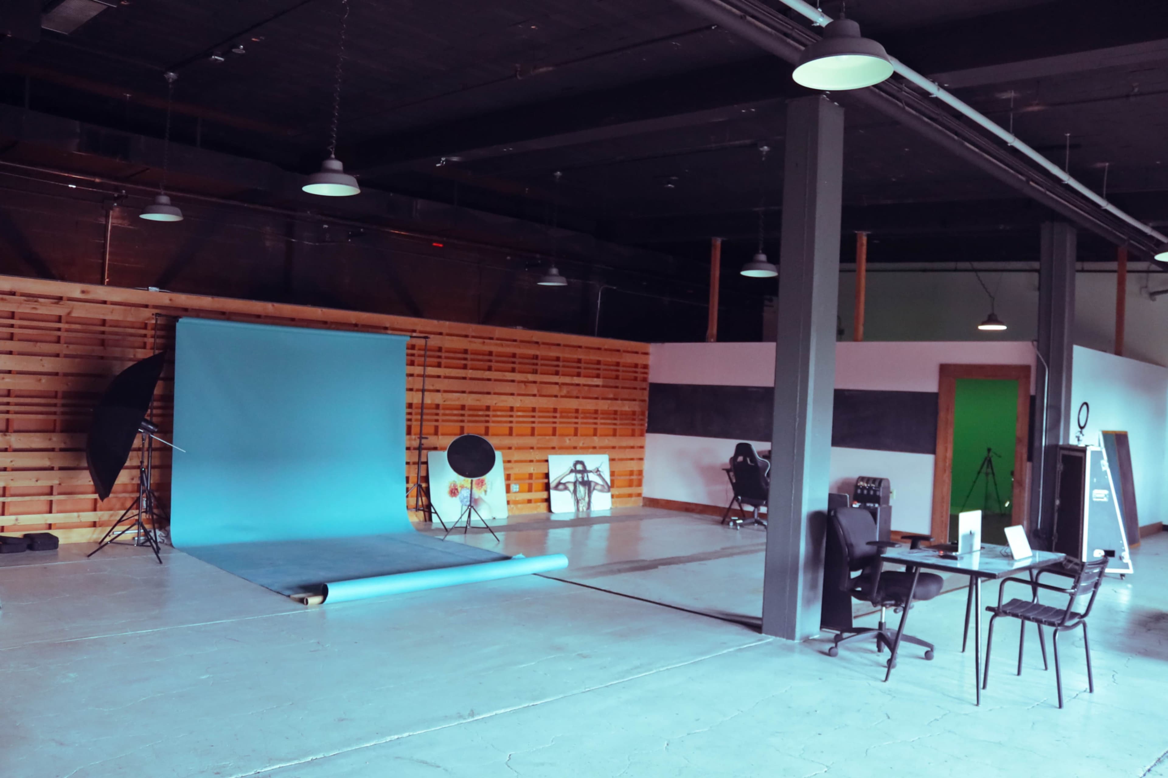 The image shows a photography studio featuring a blue backdrop, lighting equipment, and a workspace with chairs and a desk.