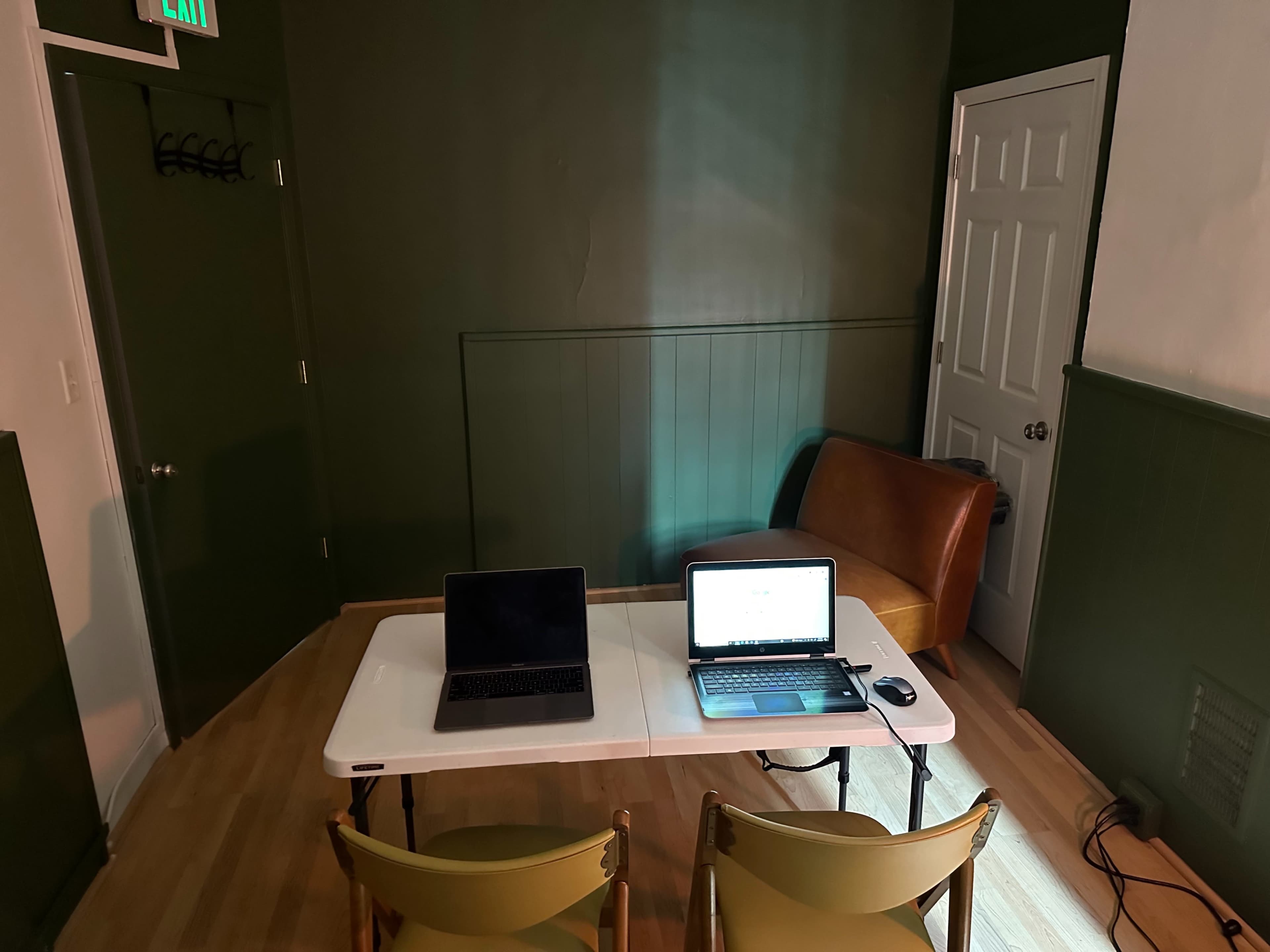 A small room features a table with two laptops, a chair, and a closed door against a dark green wall.