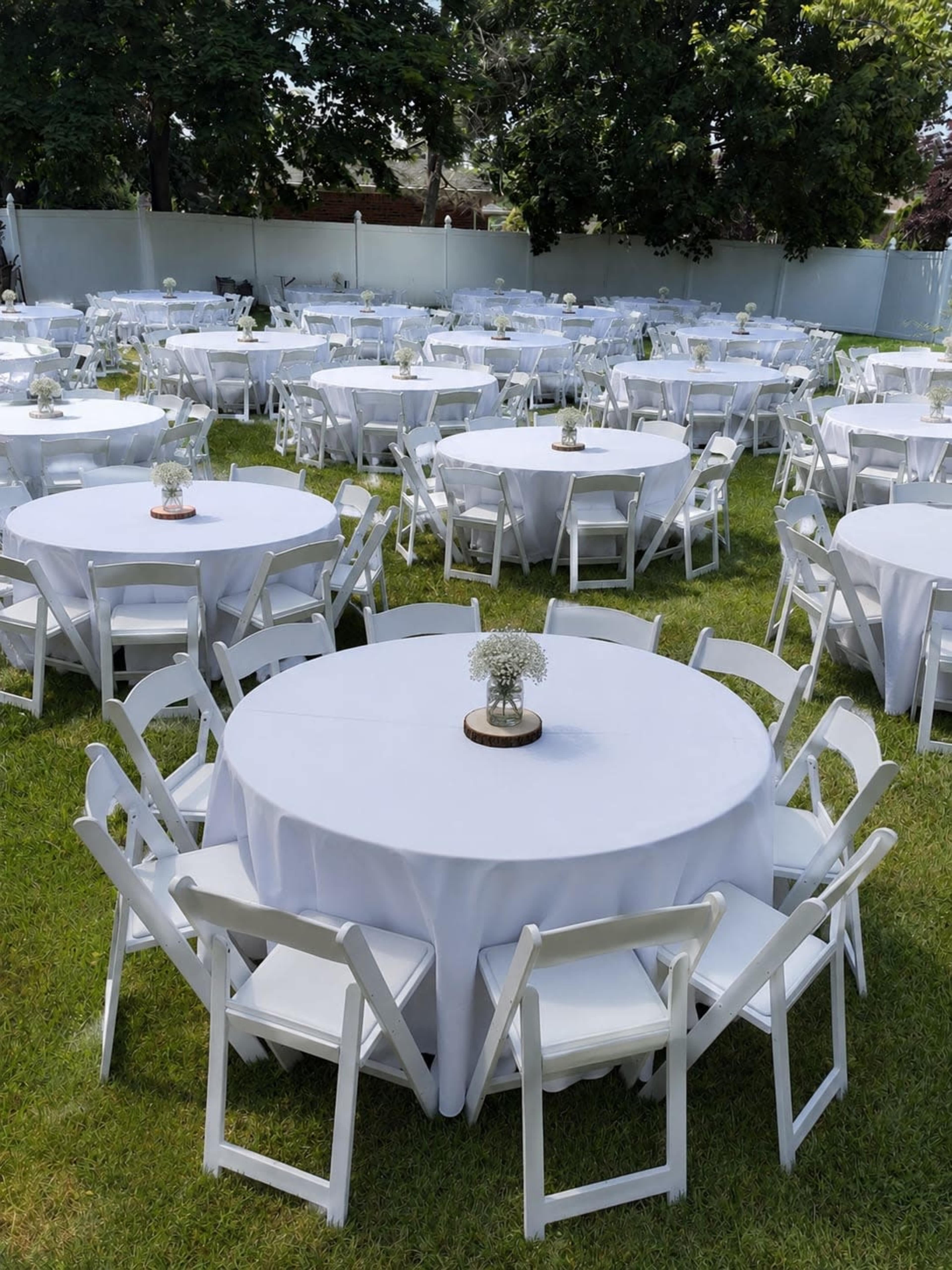 The image shows a large outdoor space filled with round tables covered in white tablecloths, each surrounded by white folding chairs and adorned with small centerpieces.