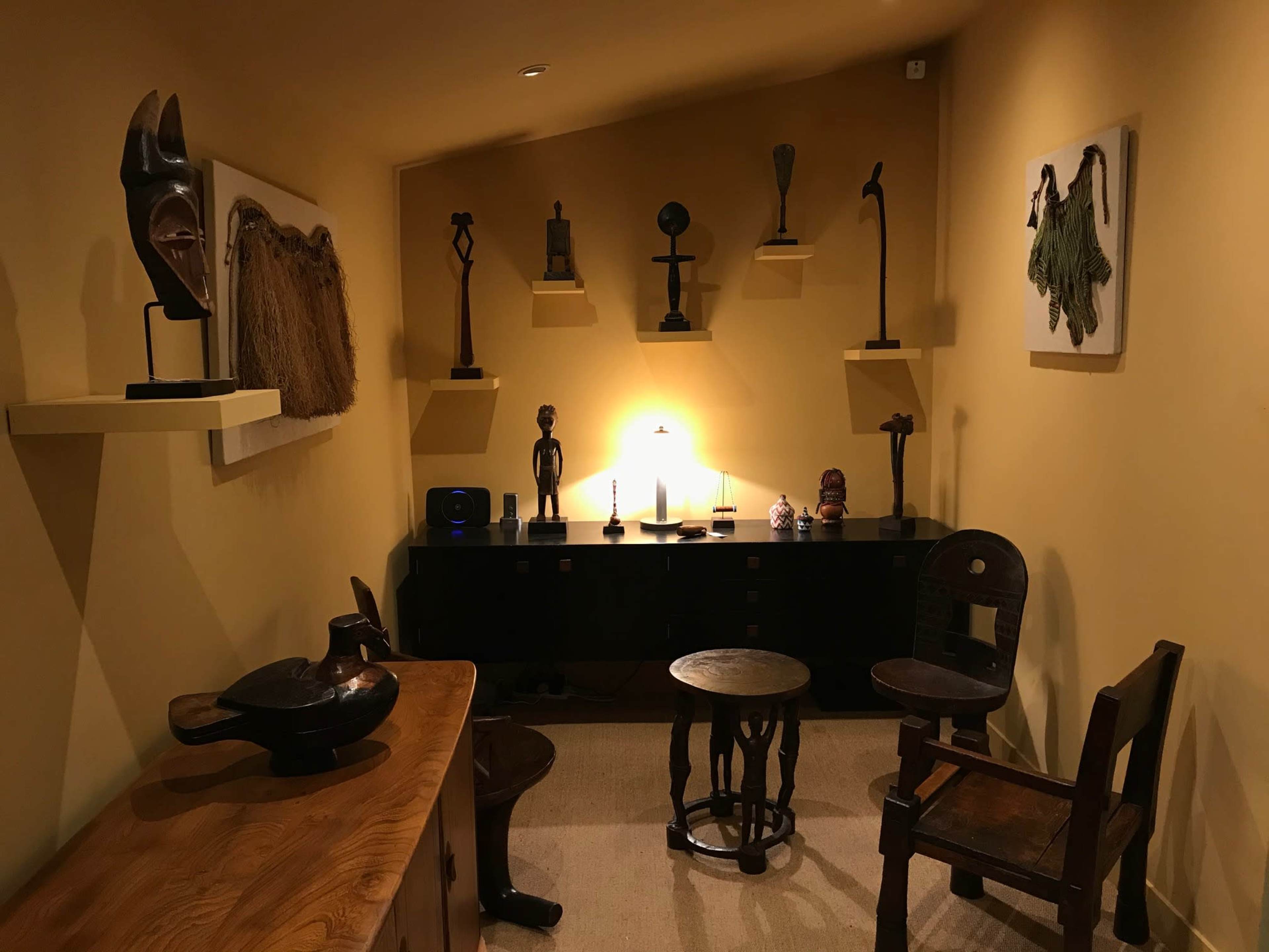 A small room with various African-style artifacts on display, including sculptures and masks, along with several wooden chairs and a low table.