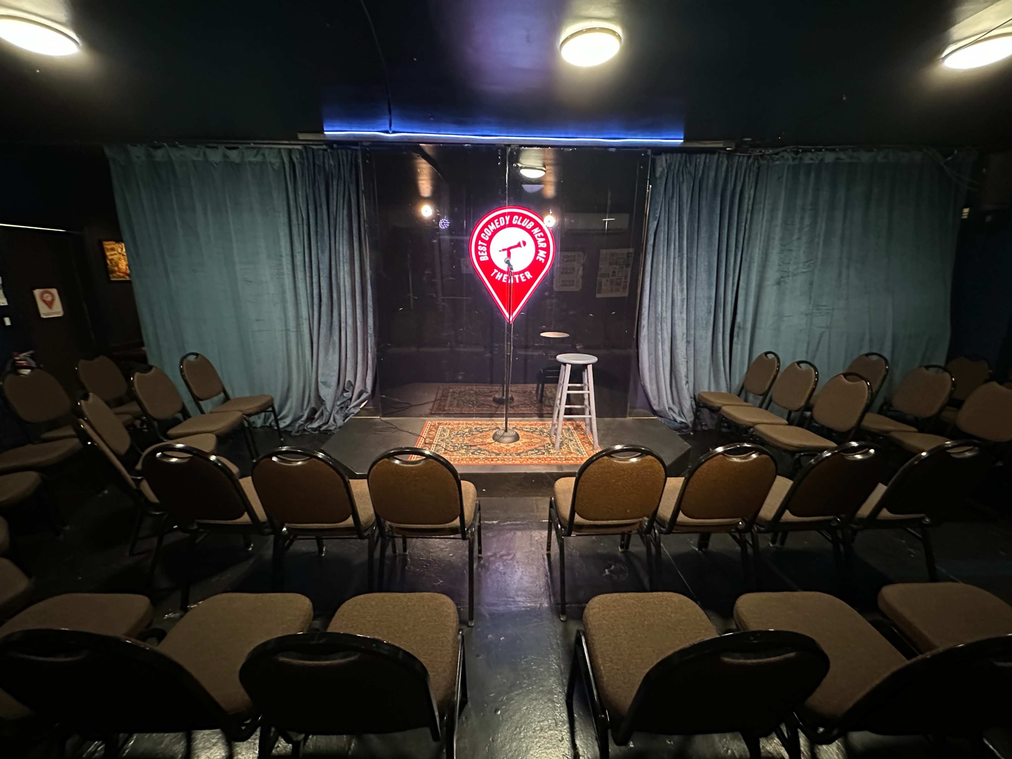 The image shows a comedy stage with a microphone stand and a stool, flanked by blue curtains, surrounded by rows of empty chairs.