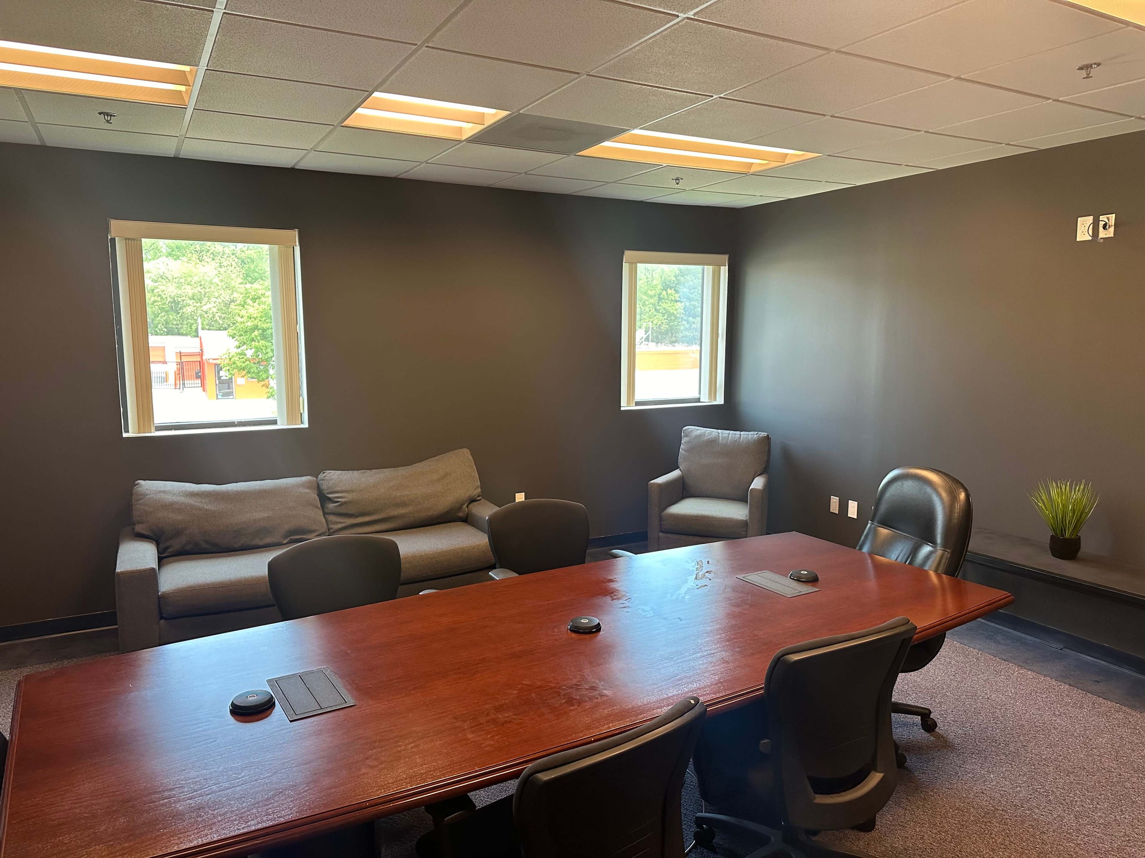 A conference room featuring a wooden table surrounded by black chairs, with a couch and an armchair against the wall and two windows letting in natural light.