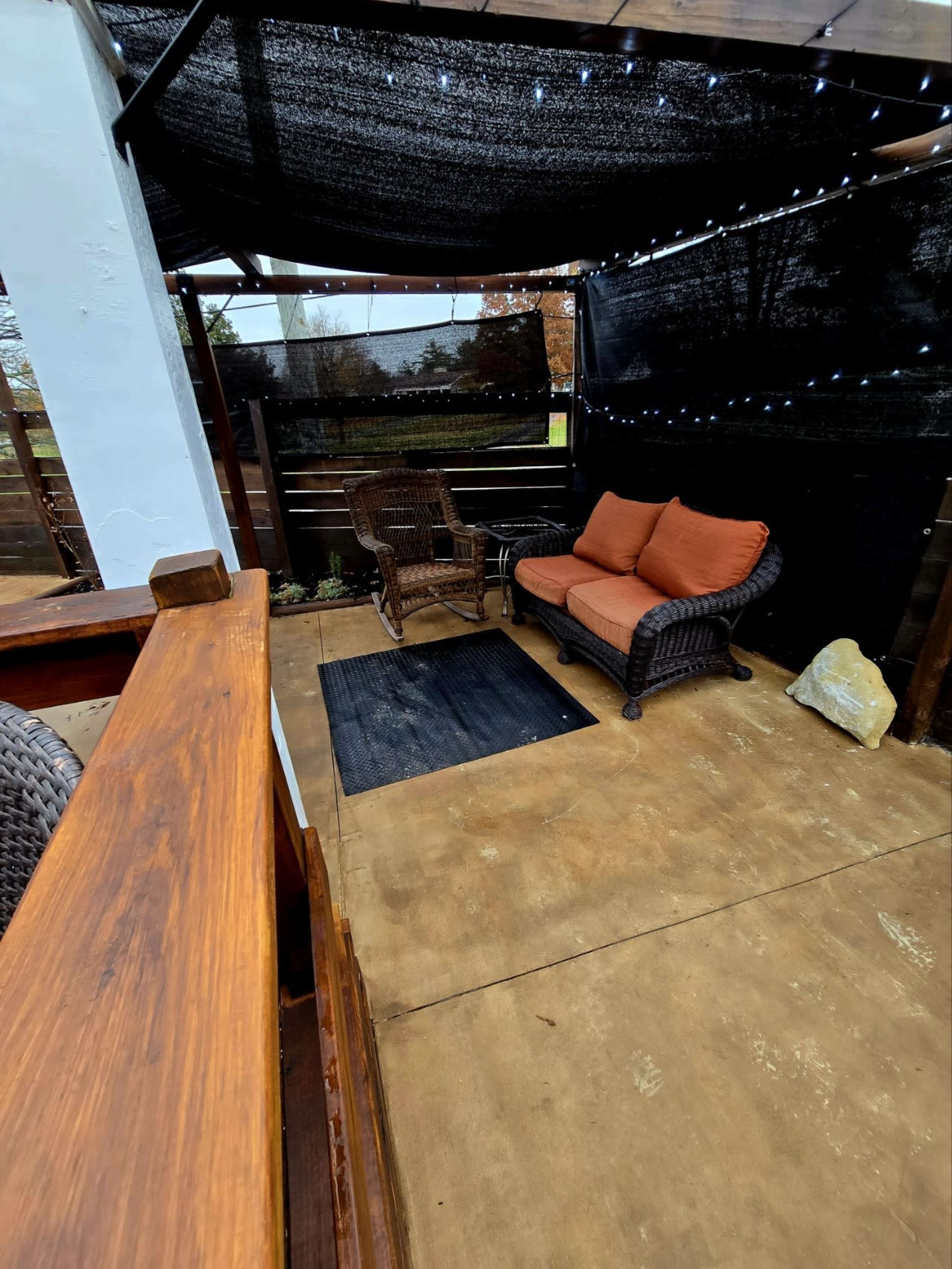 A cozy outdoor seating area features a wicker loveseat with orange cushions, set beneath a shaded canopy with string lights, on a concrete floor.