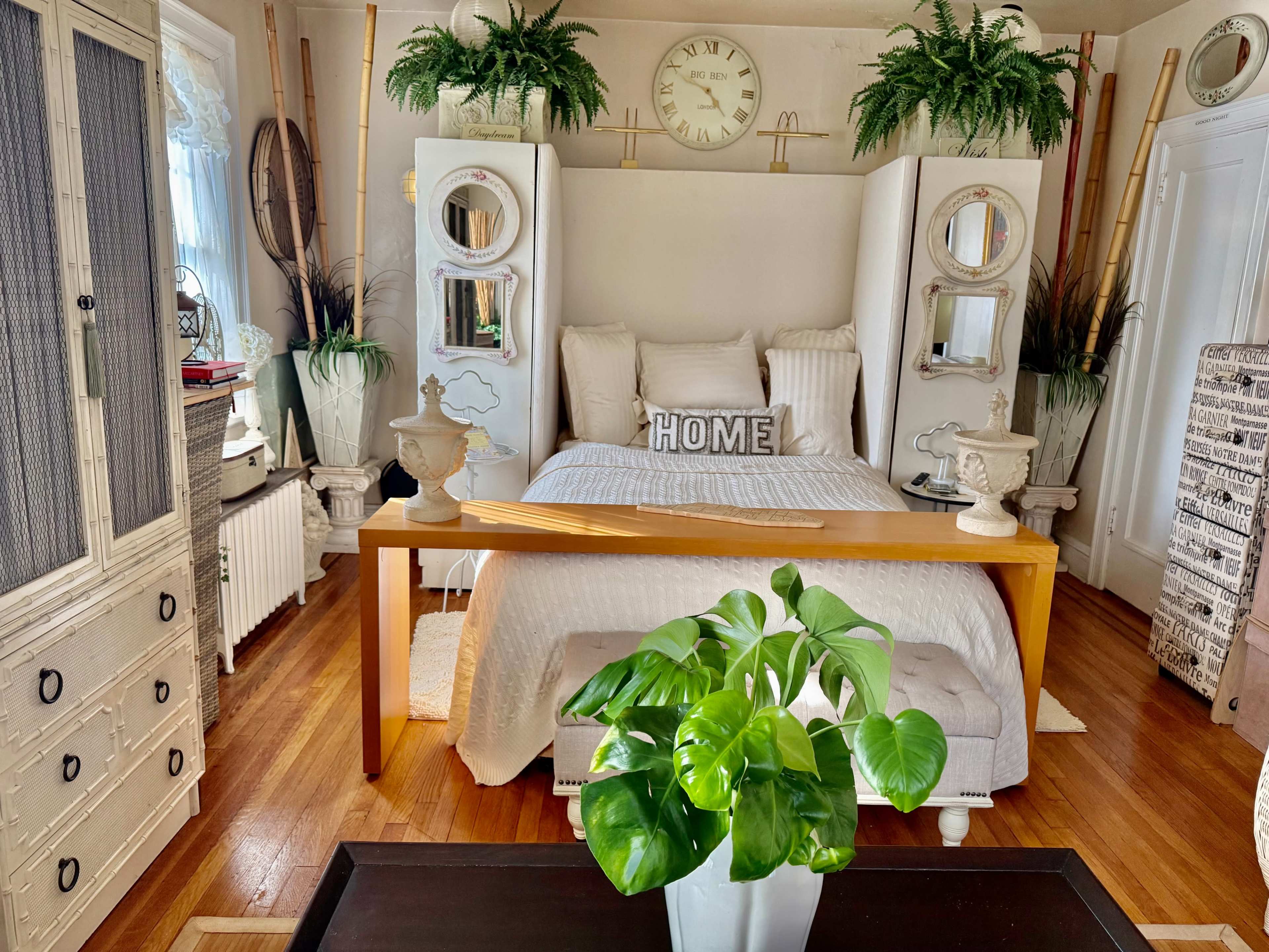 The image shows a cozy bedroom featuring a centrally placed bed with a decorative "HOME" pillow, flanked by white cabinets and surrounded by various indoor plants and furnishings.