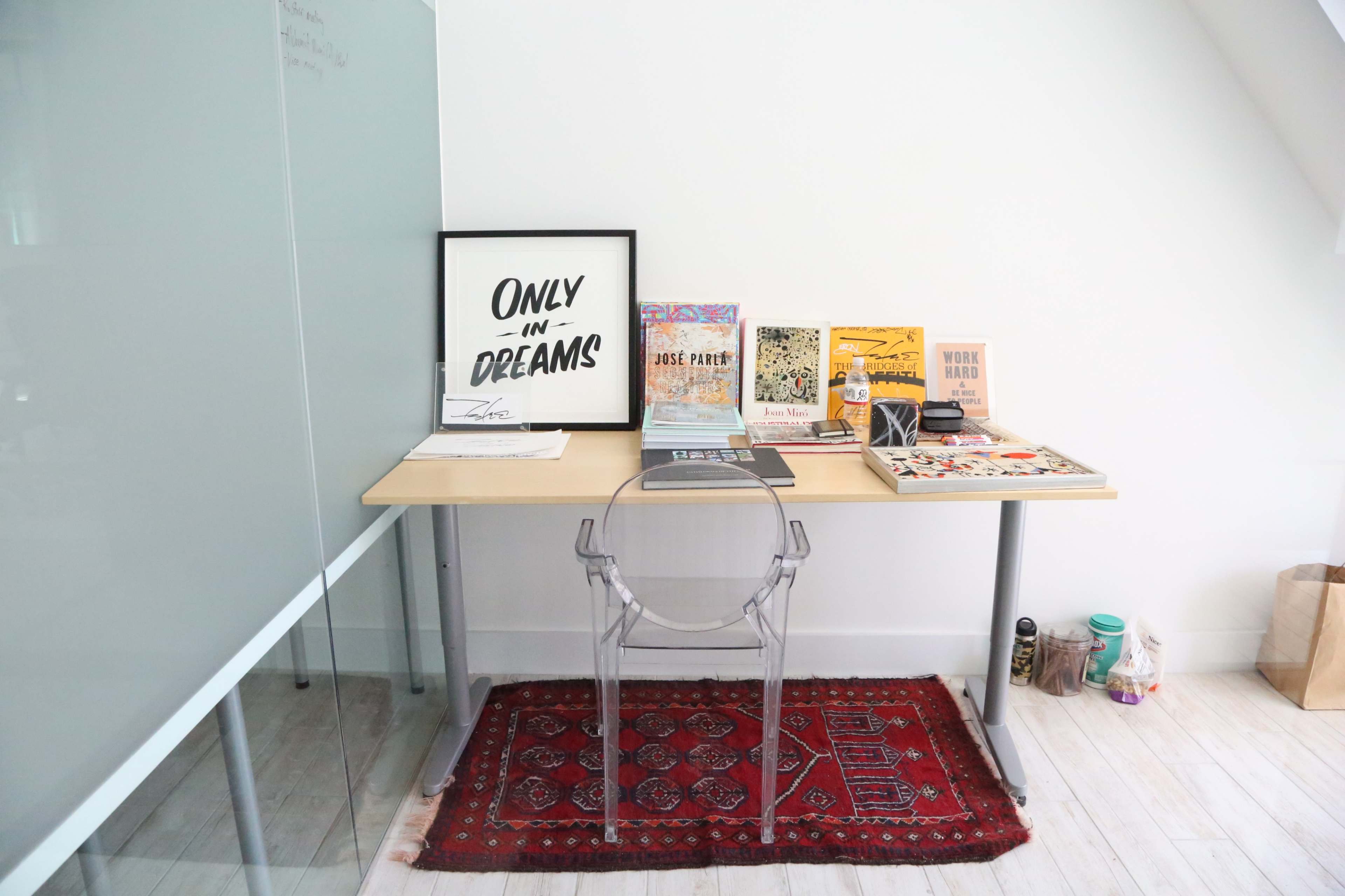 A desk with a transparent chair is positioned against a wall, featuring various books, a framed print that reads "ONLY IN DREAMS," and a colorful rug underneath.