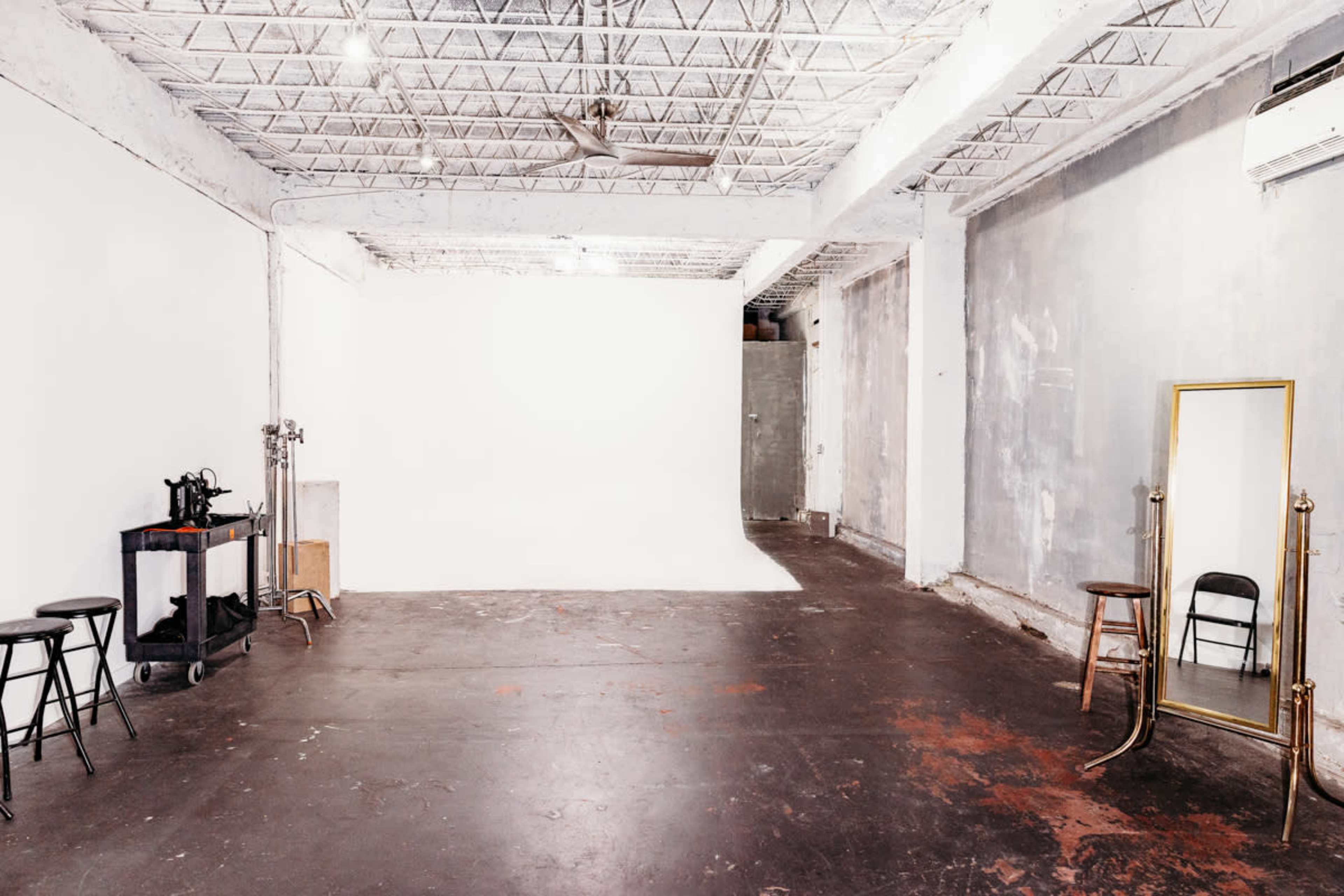 A spacious studio with exposed concrete walls, a plain white backdrop, and a few stools along with a mirror and a cart.