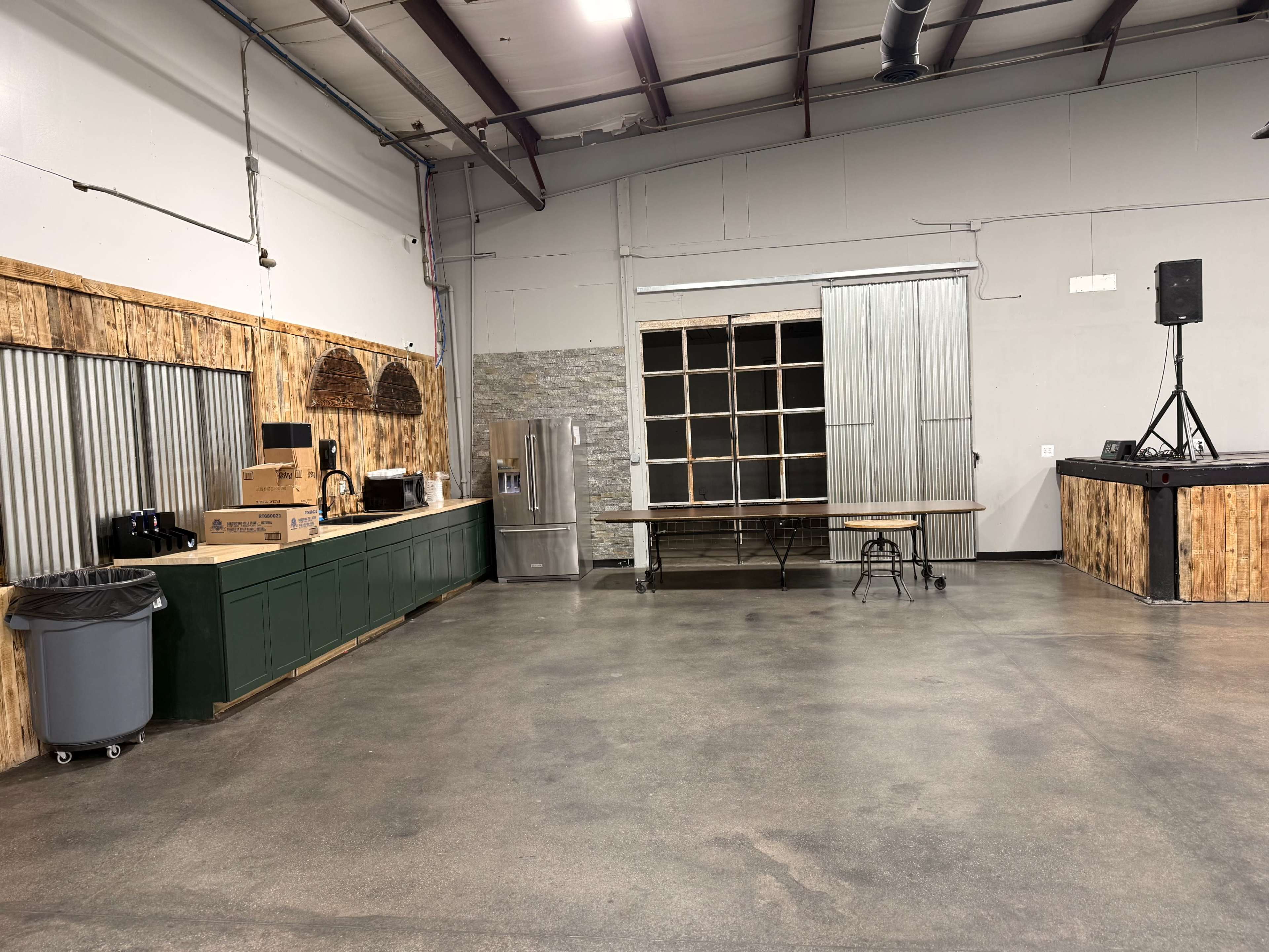 The image shows an industrial-style open space with a kitchen area featuring cabinets, a refrigerator, and a table, along with a stage set up in a corner.