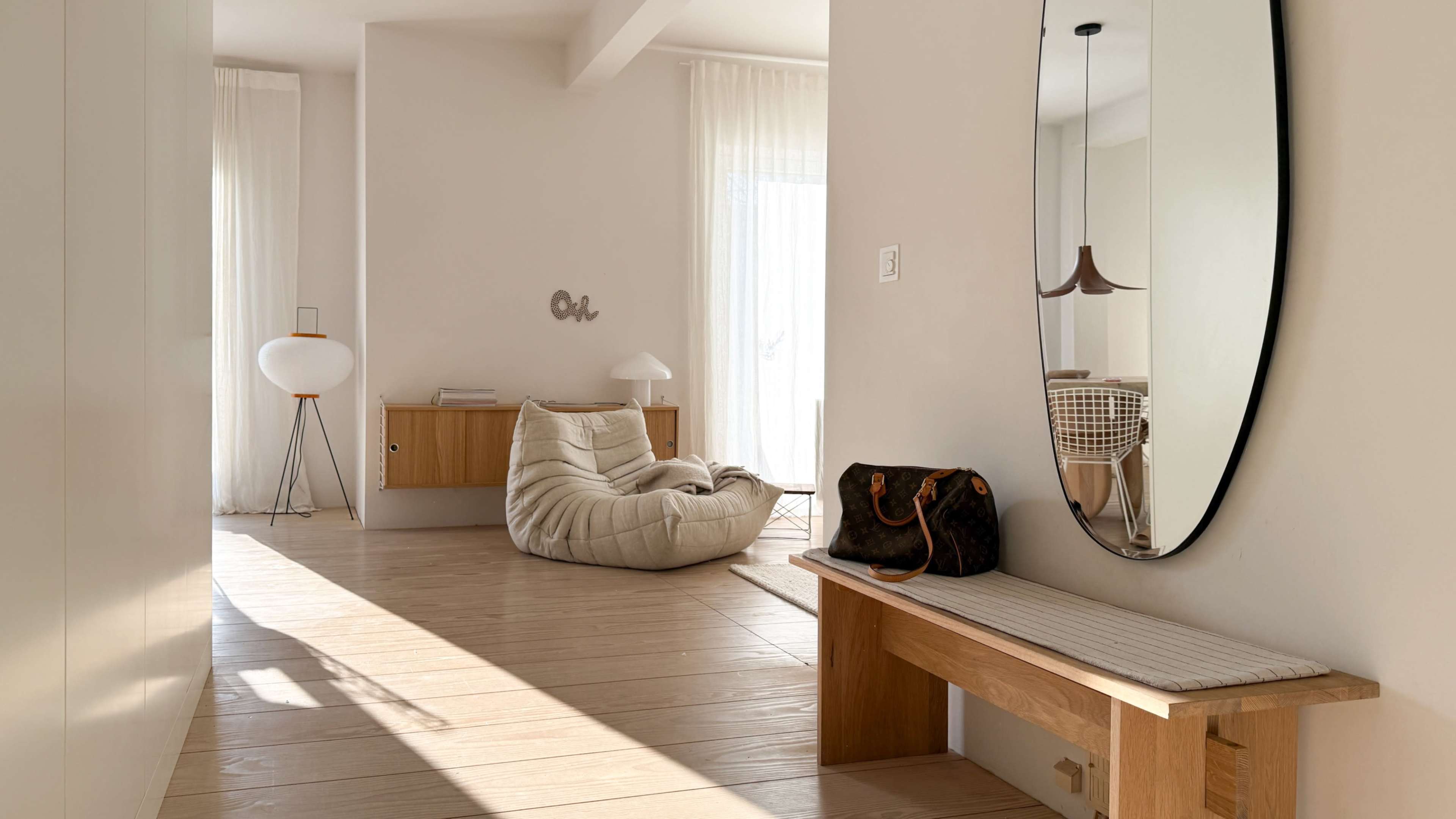 The image shows a modern, minimalist interior space featuring a wooden bench, a large mirror, a bean bag chair, and soft lighting from large windows.