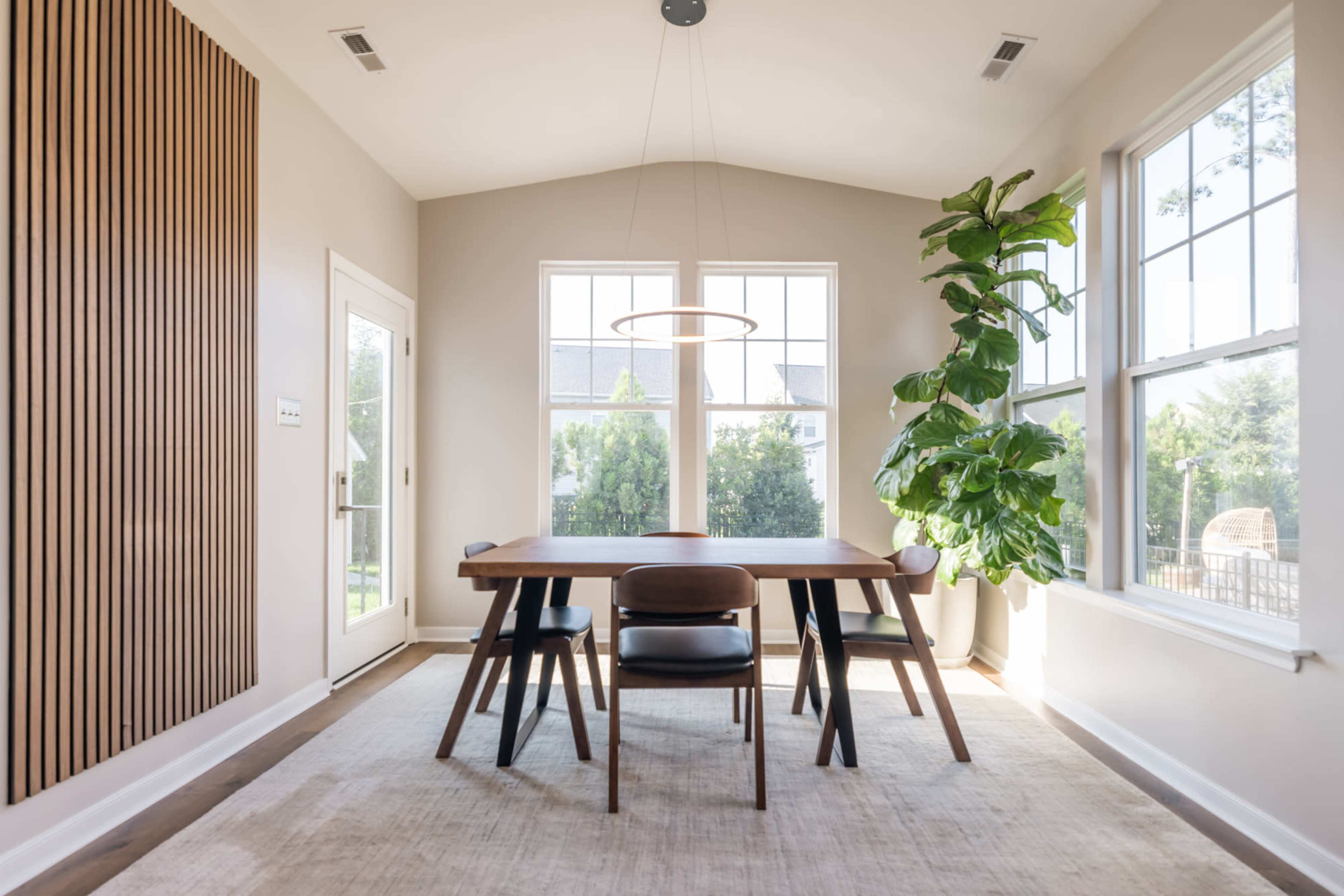 A dining area features a wooden table with four chairs, a large pendant light, and a tall plant, surrounded by windows that let in abundant natural light.