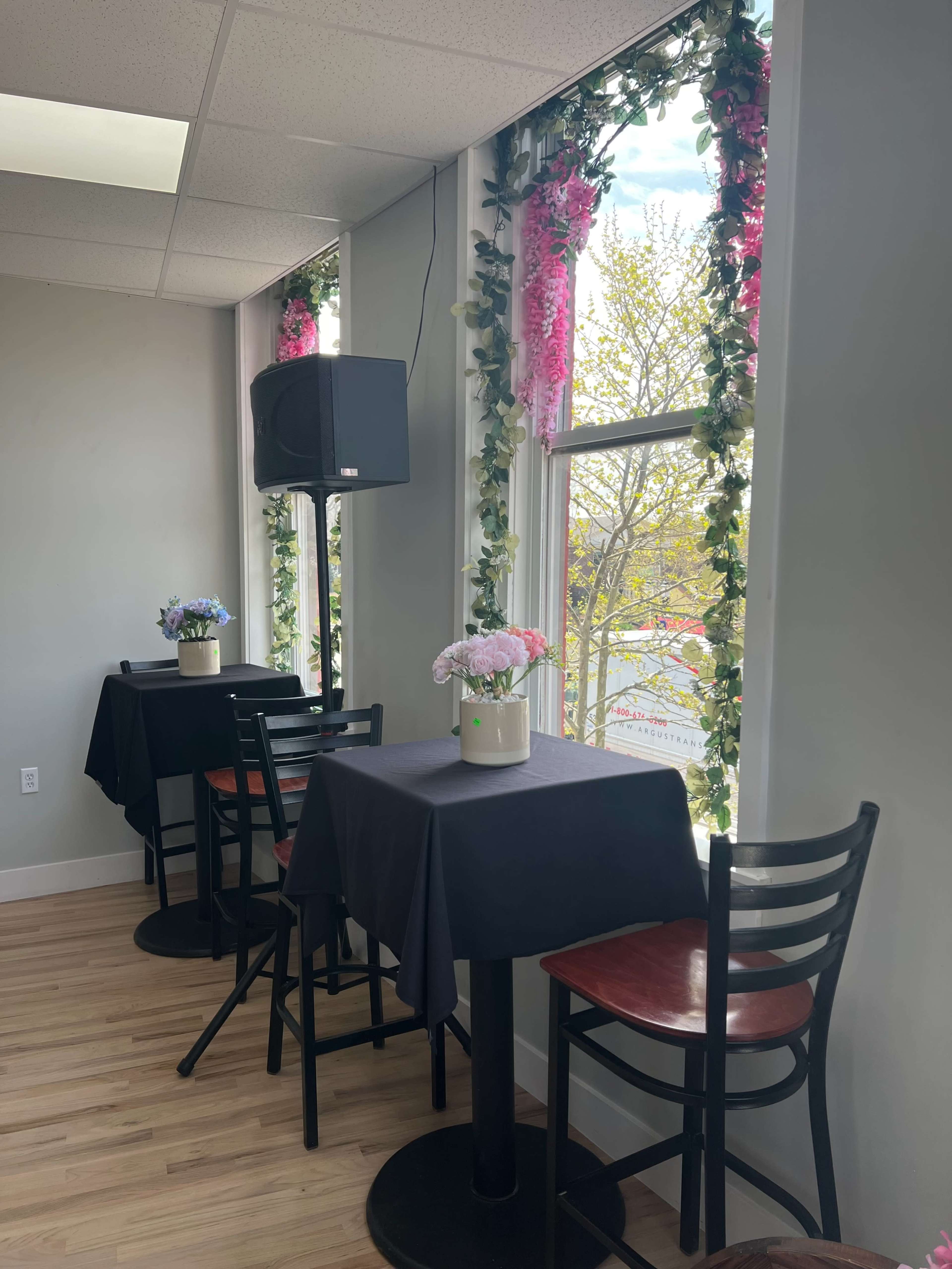 The image shows a small dining area with black tables and chairs, accented by flower arrangements and greenery near the windows.
