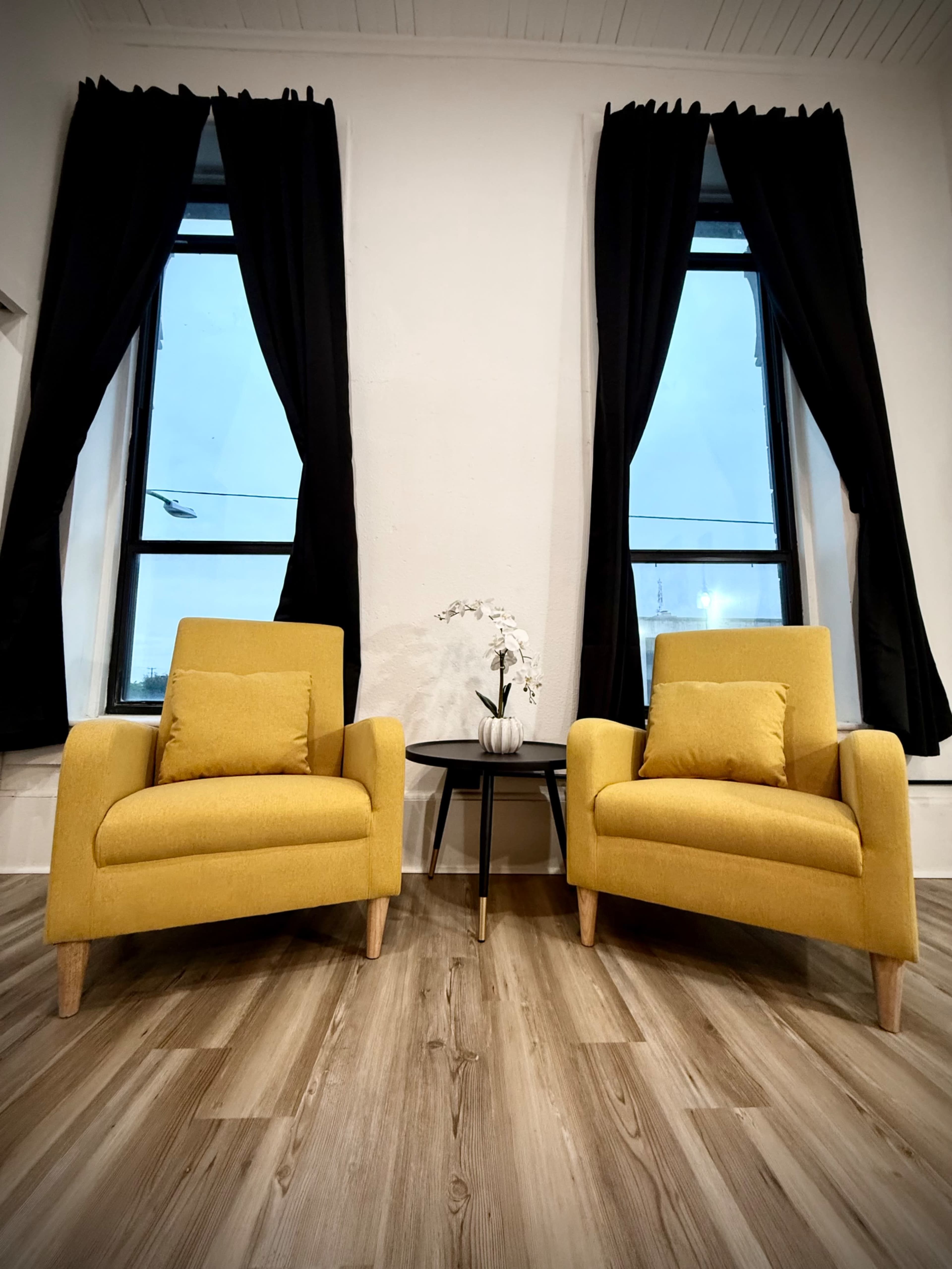 Two yellow armchairs are positioned in front of large windows with black curtains, alongside a small round table decorated with a white vase.