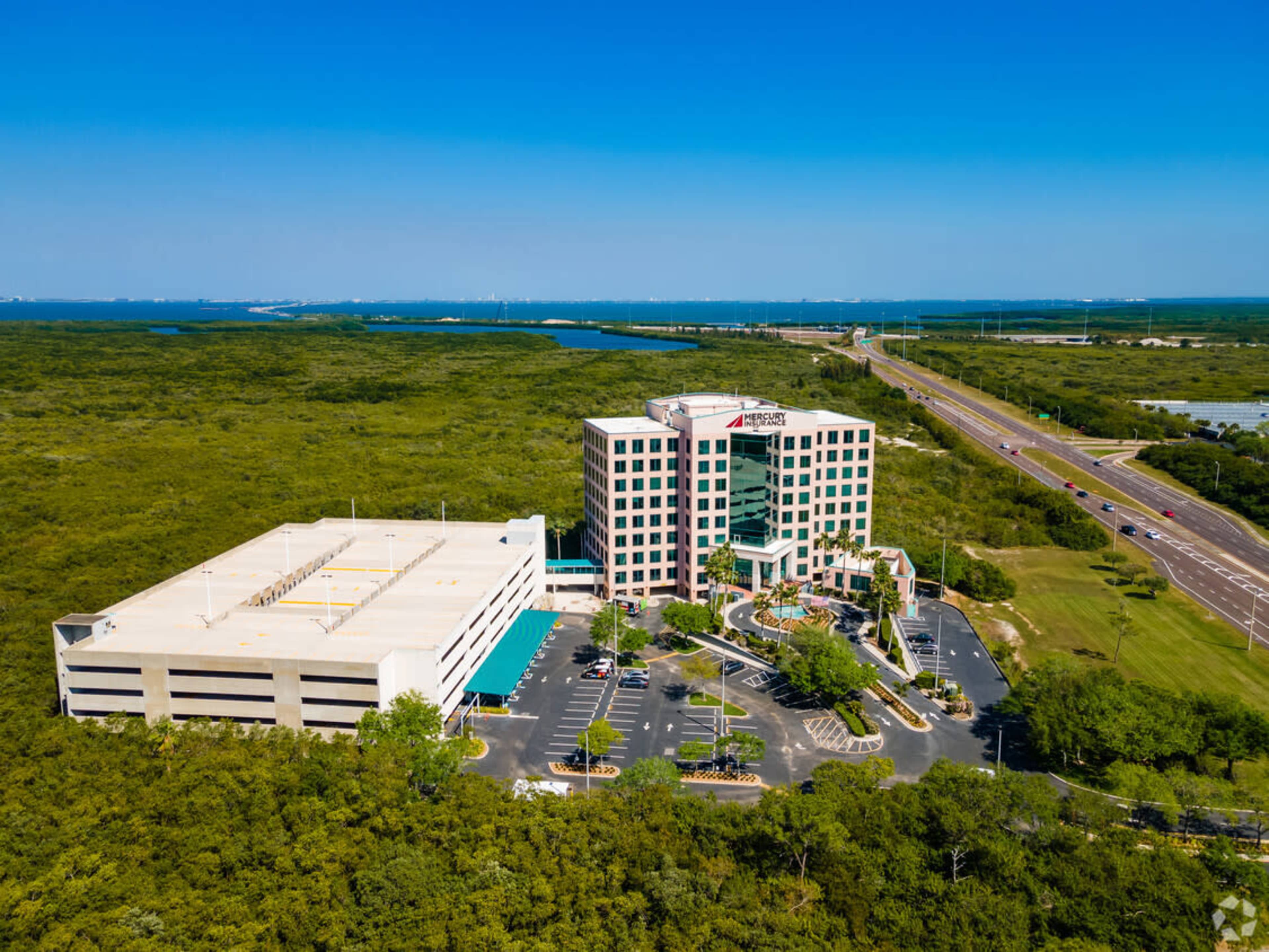 A multi-story hotel building with a parking garage is situated near a highway and surrounded by greenery and water.