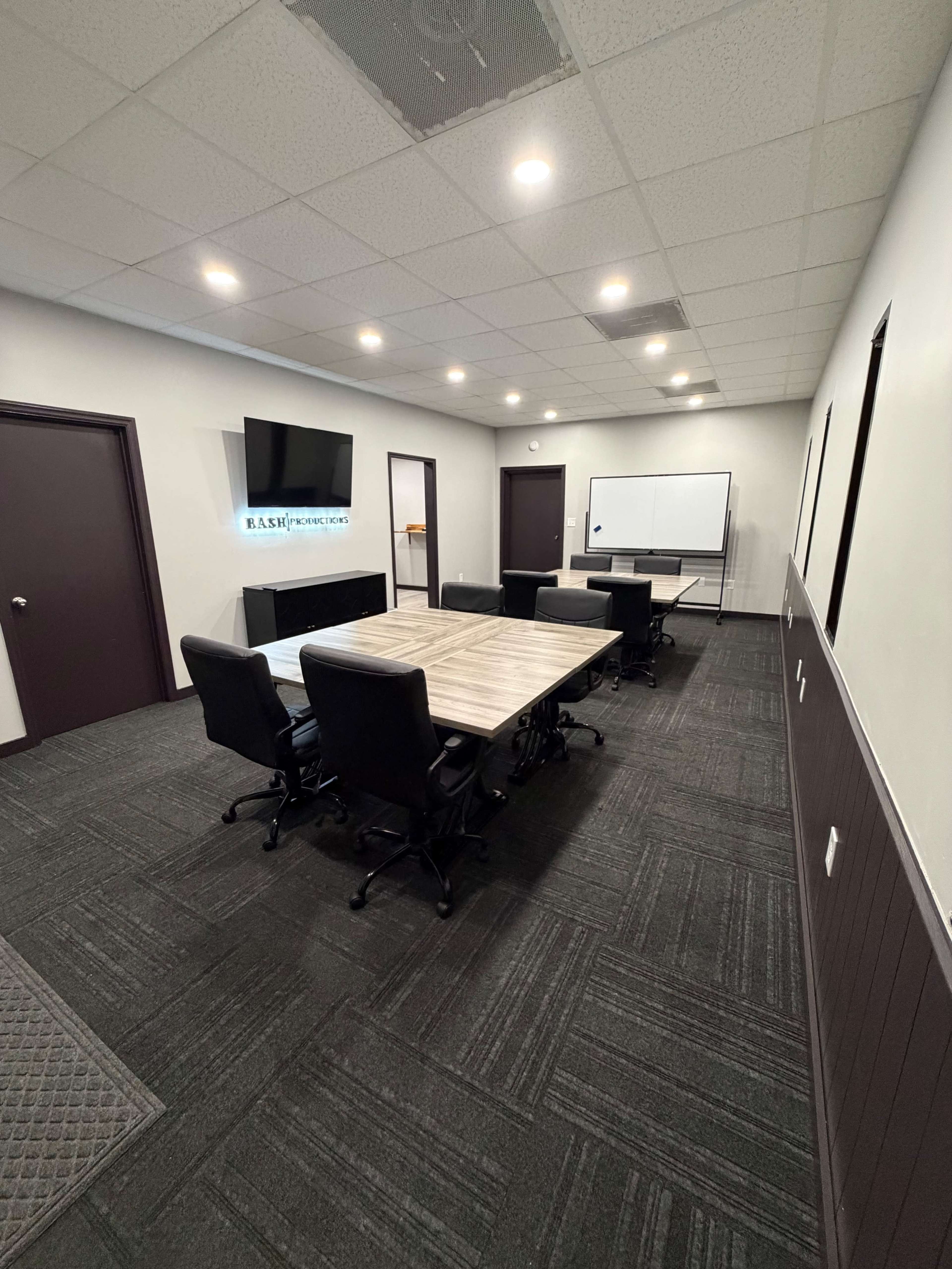 The image shows a modern conference room equipped with a large table, multiple chairs, a wall-mounted TV, and a whiteboard.
