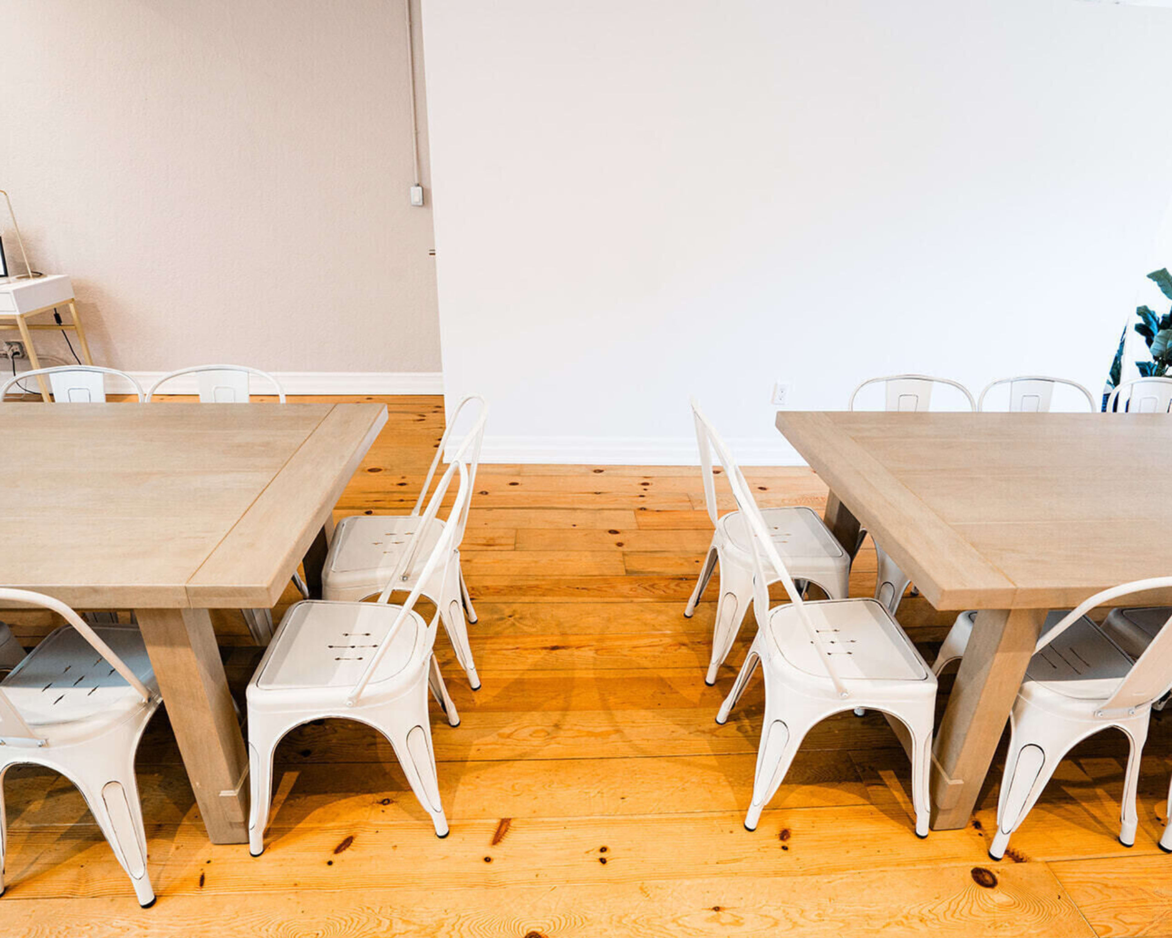 Two wooden tables are positioned facing each other on a wooden floor, surrounded by white metal chairs.
