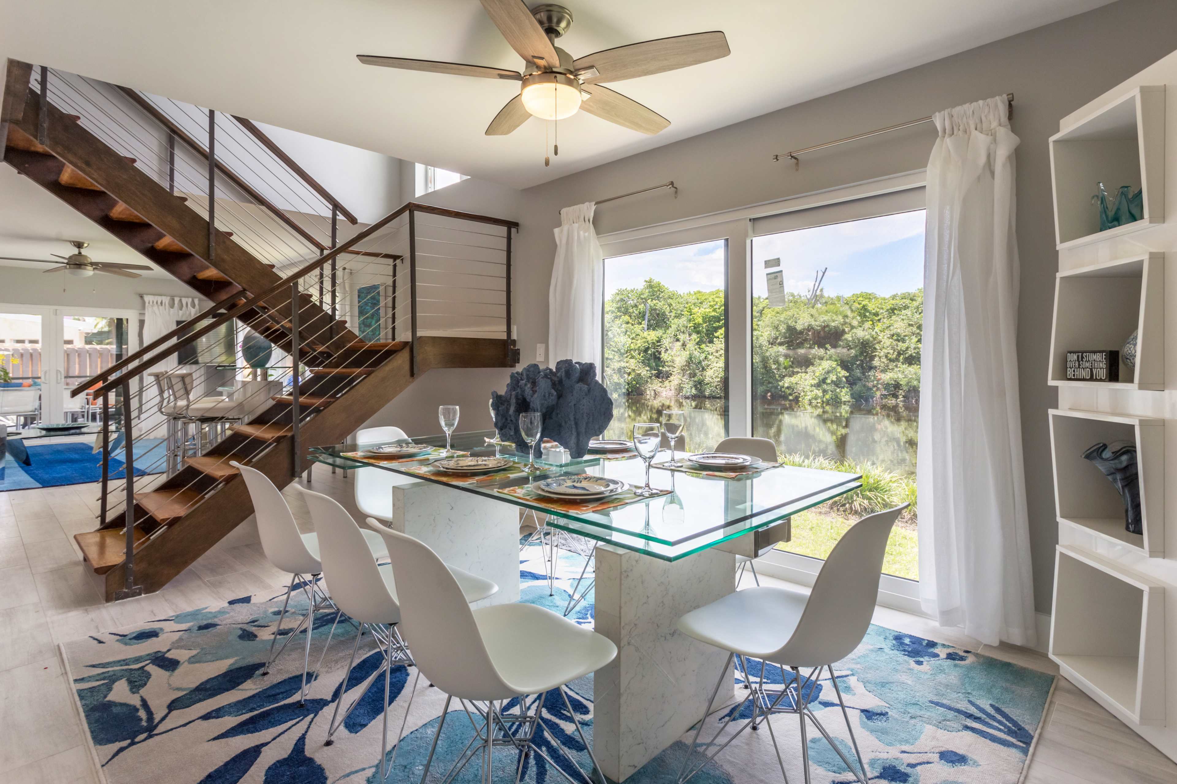 The image shows a modern dining area with a glass table set for dinner, surrounded by white chairs, and illuminated by natural light from large windows overlooking greenery.