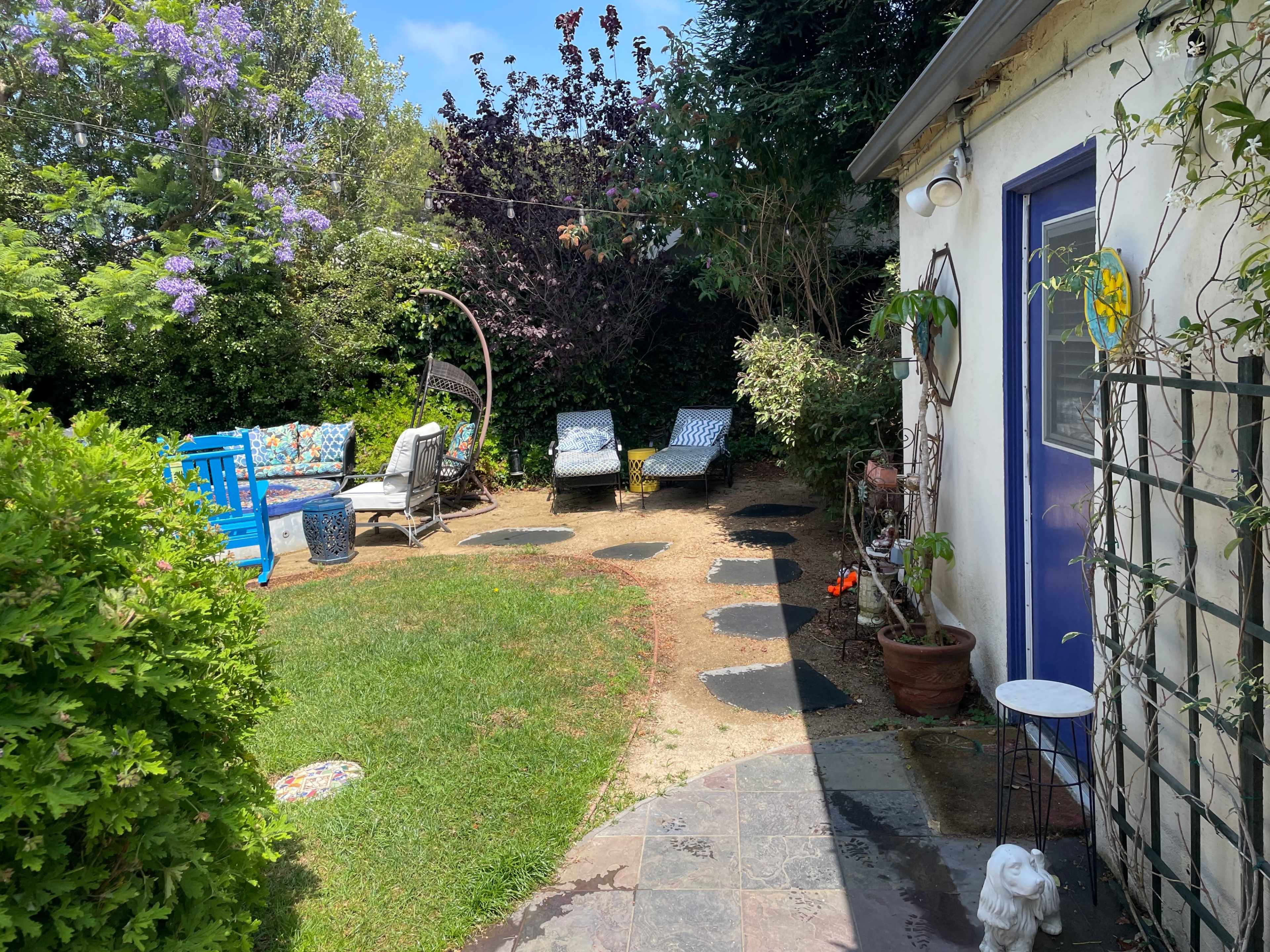The image shows a backyard with a small lawn, lounge chairs, a blue table, and a wall covered in greenery.