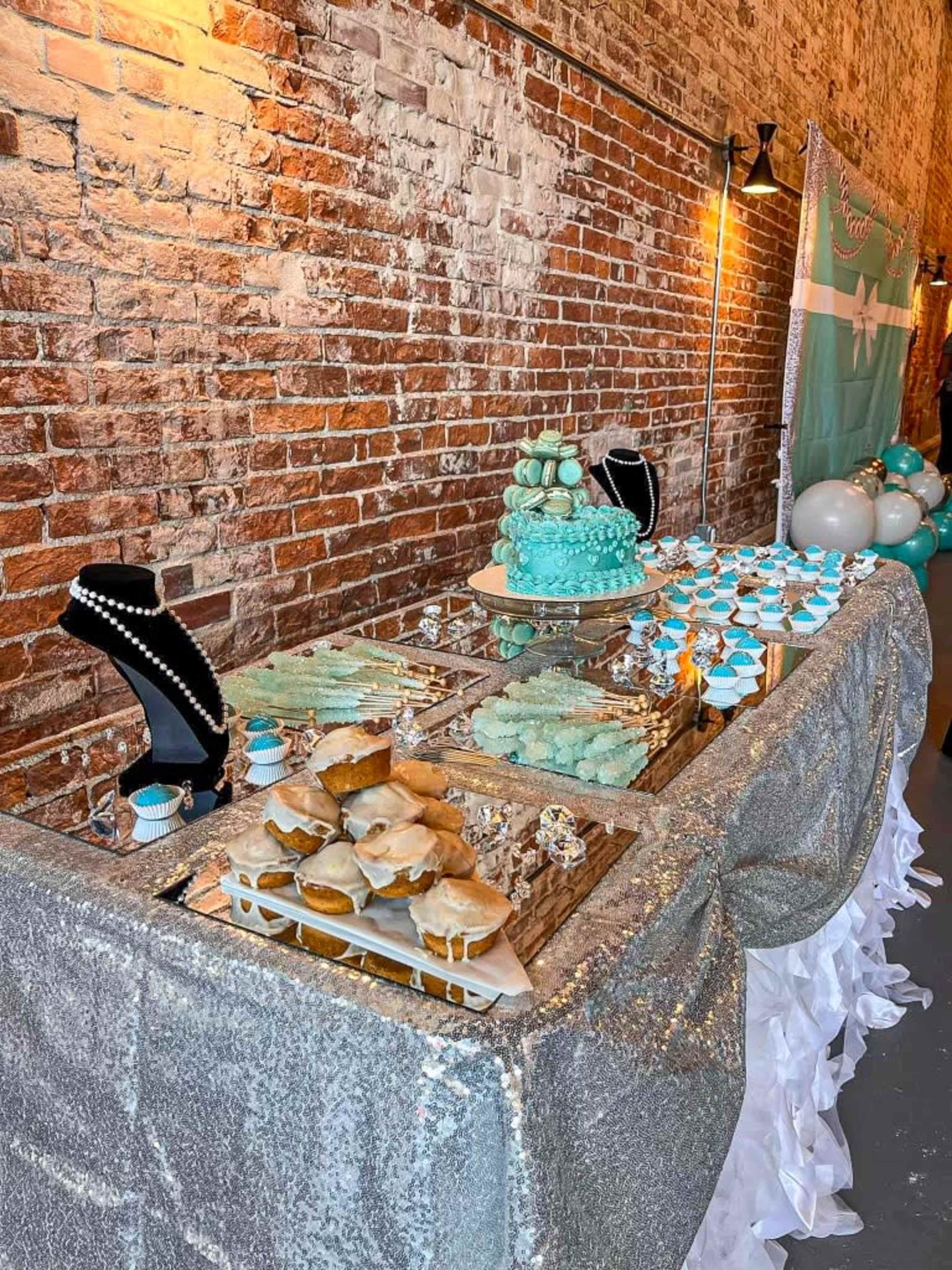 A dessert table features a turquoise cake, cupcakes, and cookies displayed under glass cloches, set against a rustic brick wall.
