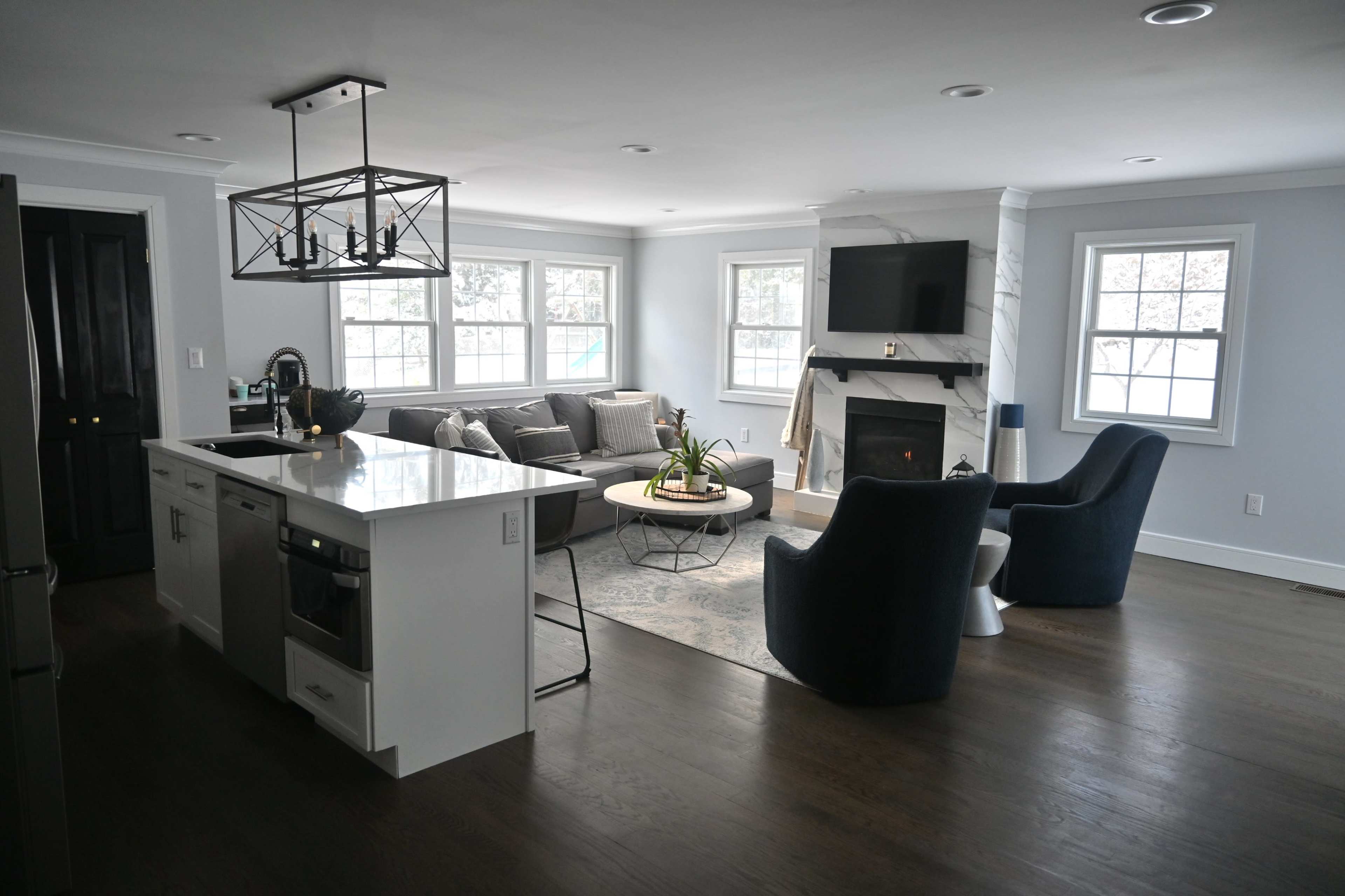 A modern living room with a light gray color scheme, featuring a sectional sofa, two blue chairs, a coffee table, and a kitchen island in the foreground.