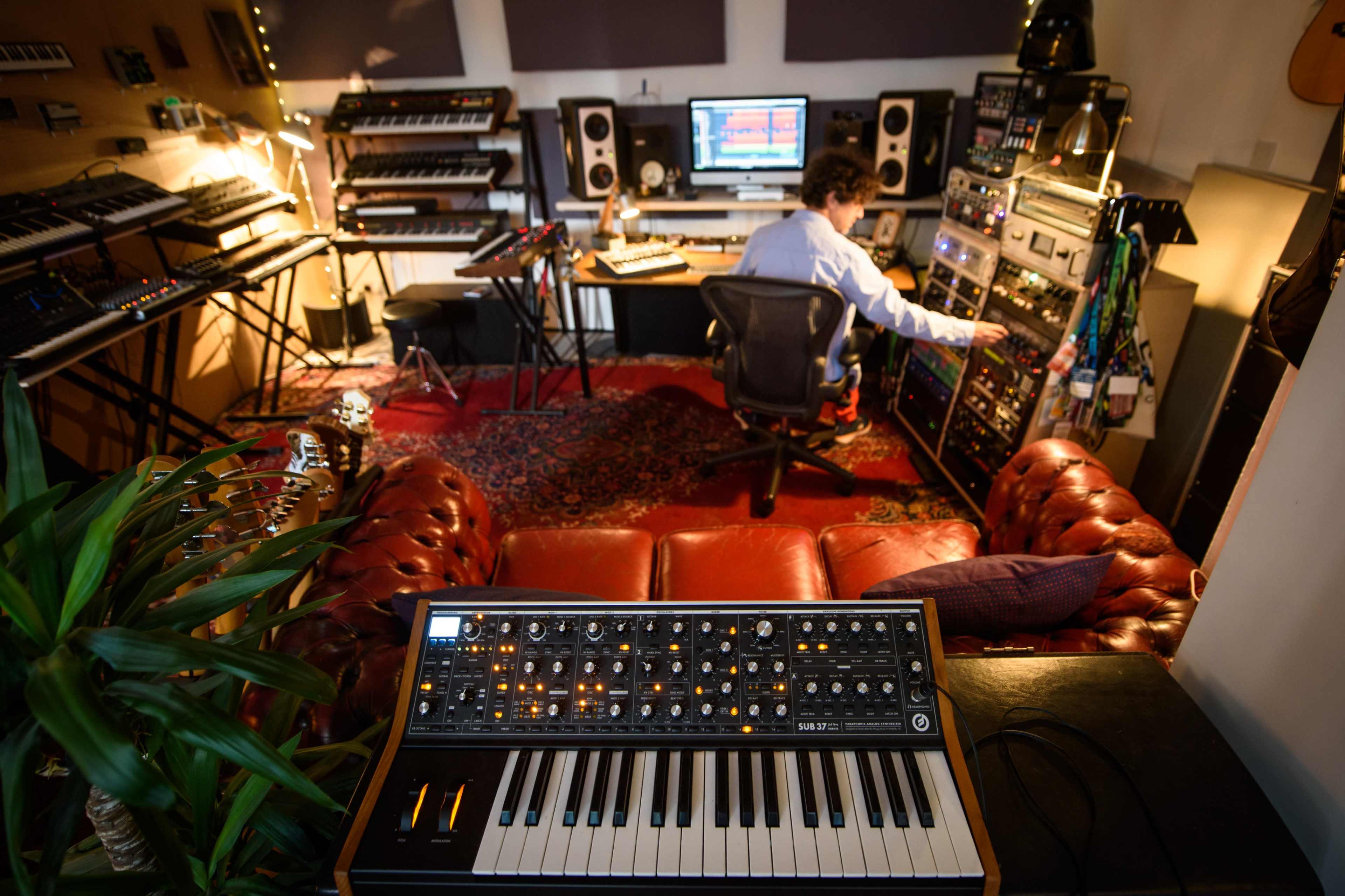 A person is seated in a music studio, adjusting equipment while surrounded by various synthesizers and a computer setup.