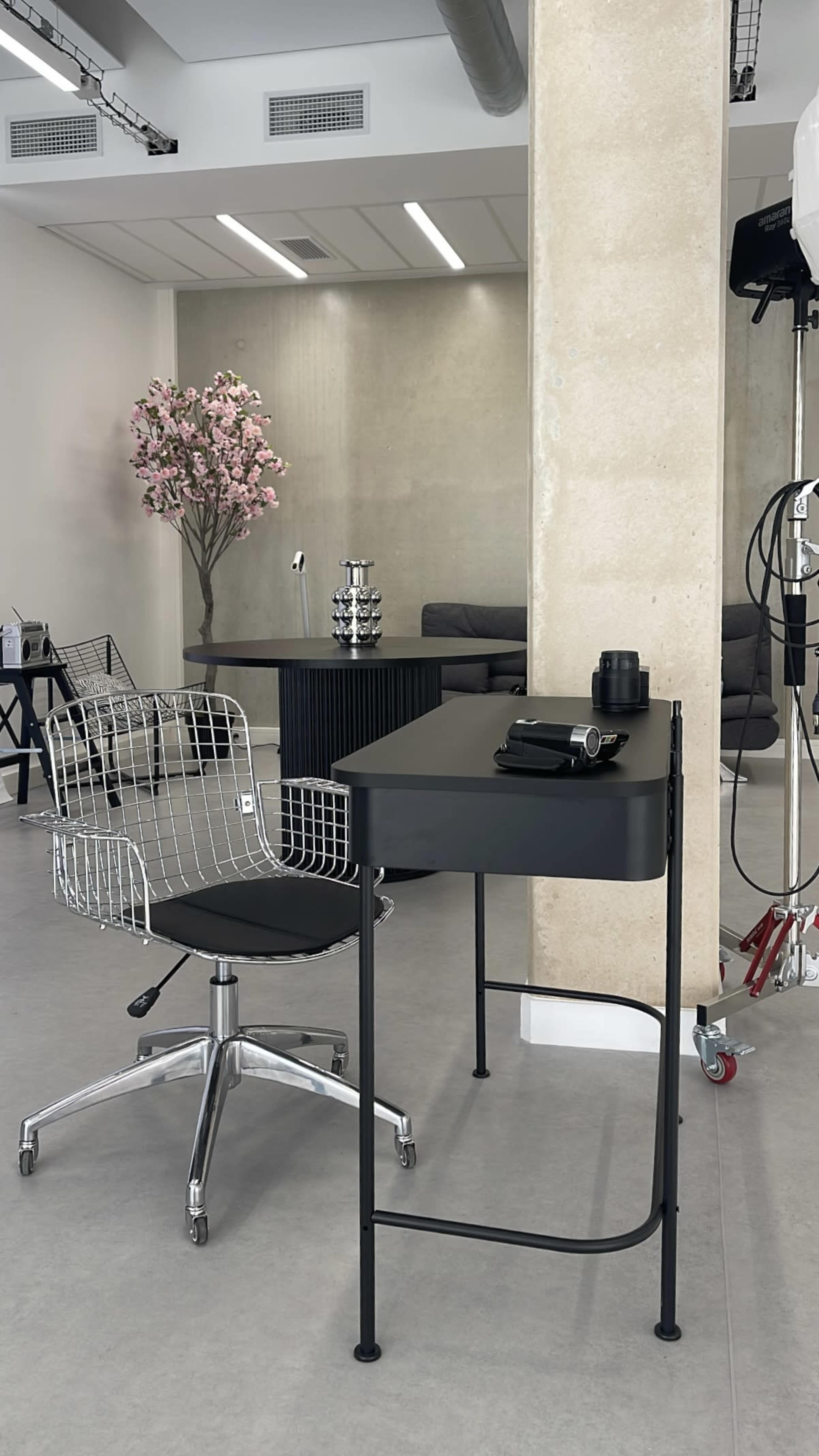 The image shows a modern workspace featuring a black desk with a chair made of wire mesh, set in a minimalistic room with light-colored walls and decorative elements like a pink flower arrangement.