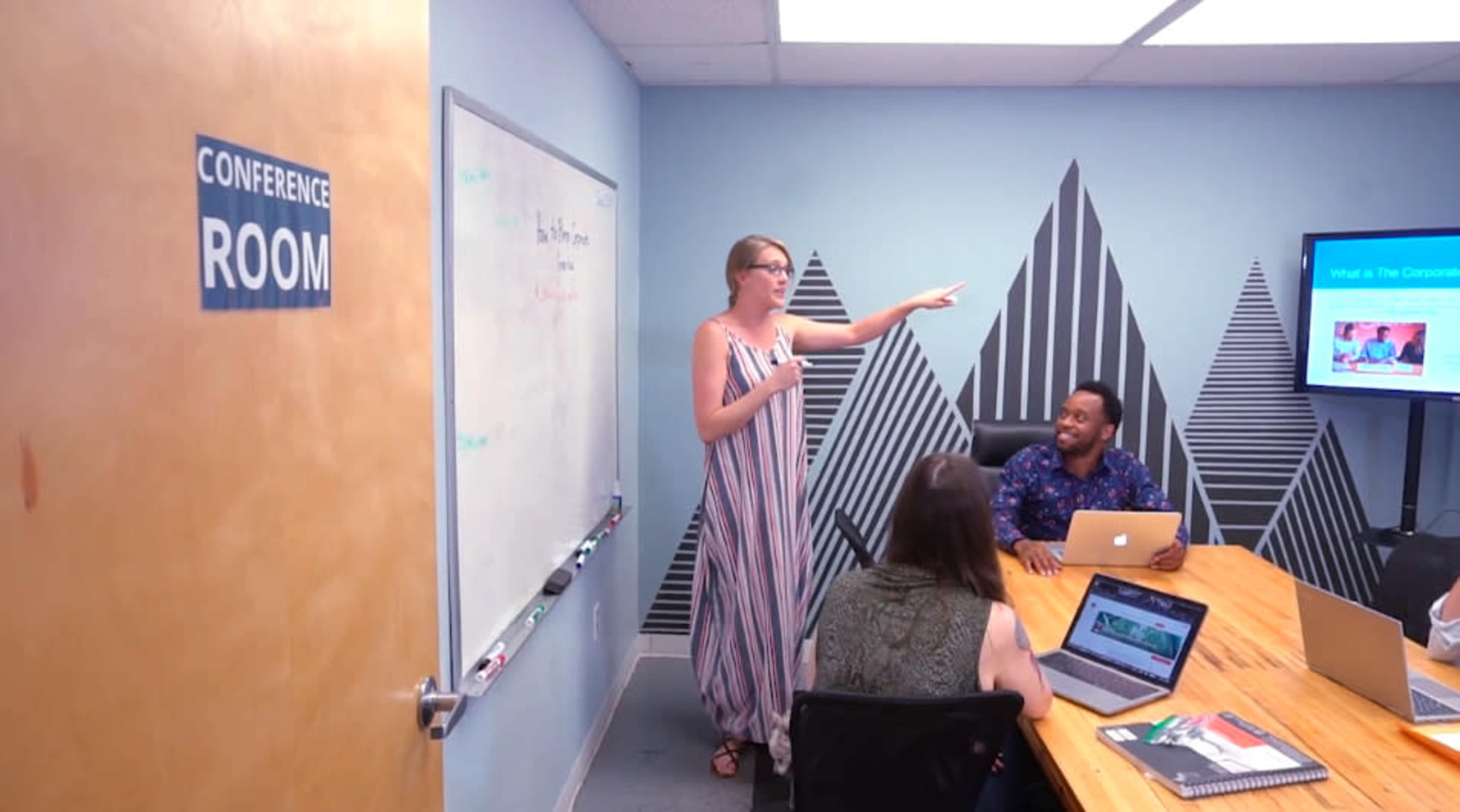 A woman gestures while presenting in a conference room where three people are seated at a table with laptops.