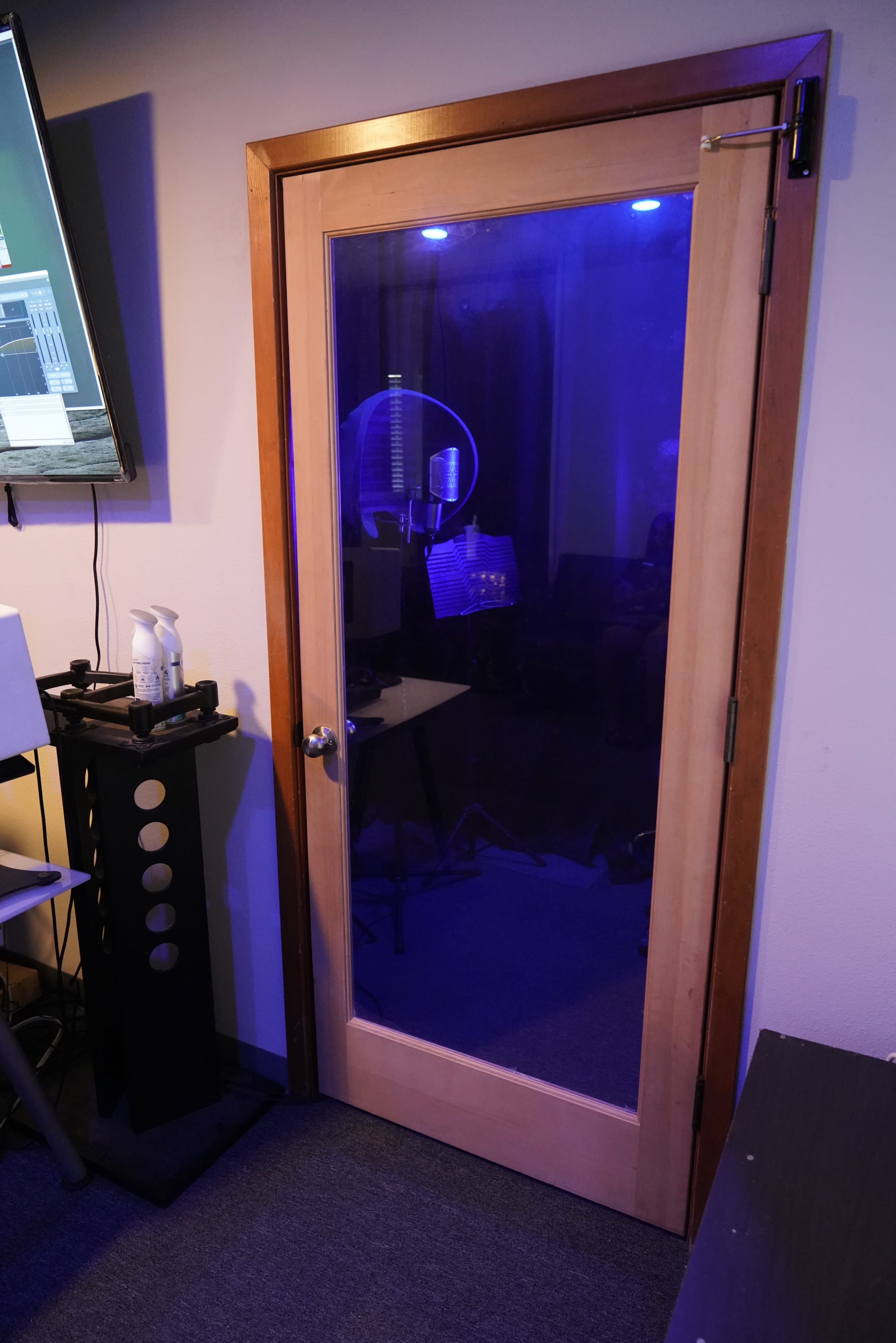 A wooden-framed glass door leading into a room with blue ambient lighting, likely a recording studio.