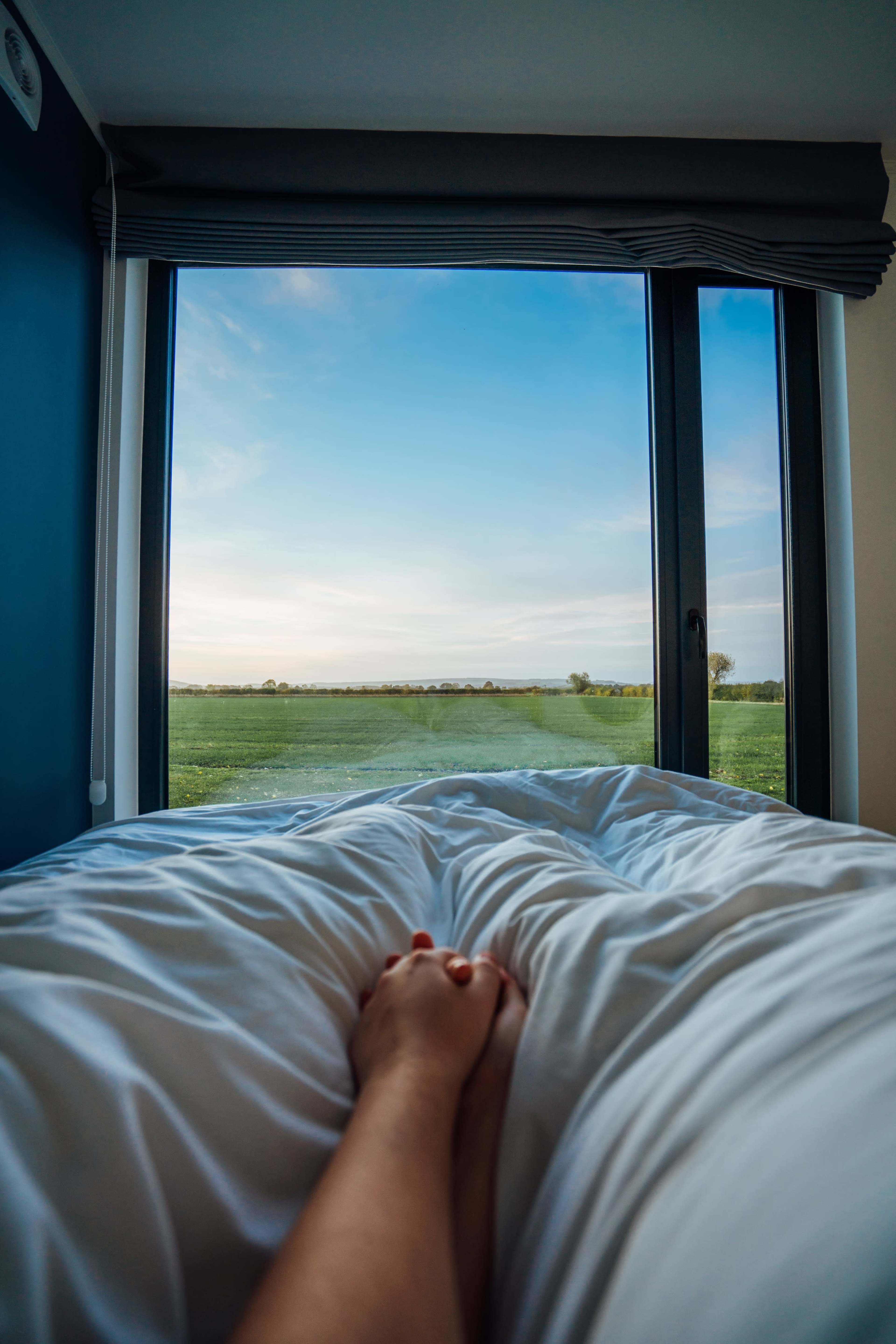 A person is holding a bed sheet while lying in bed with a view of a green field and blue sky visible through a large window.