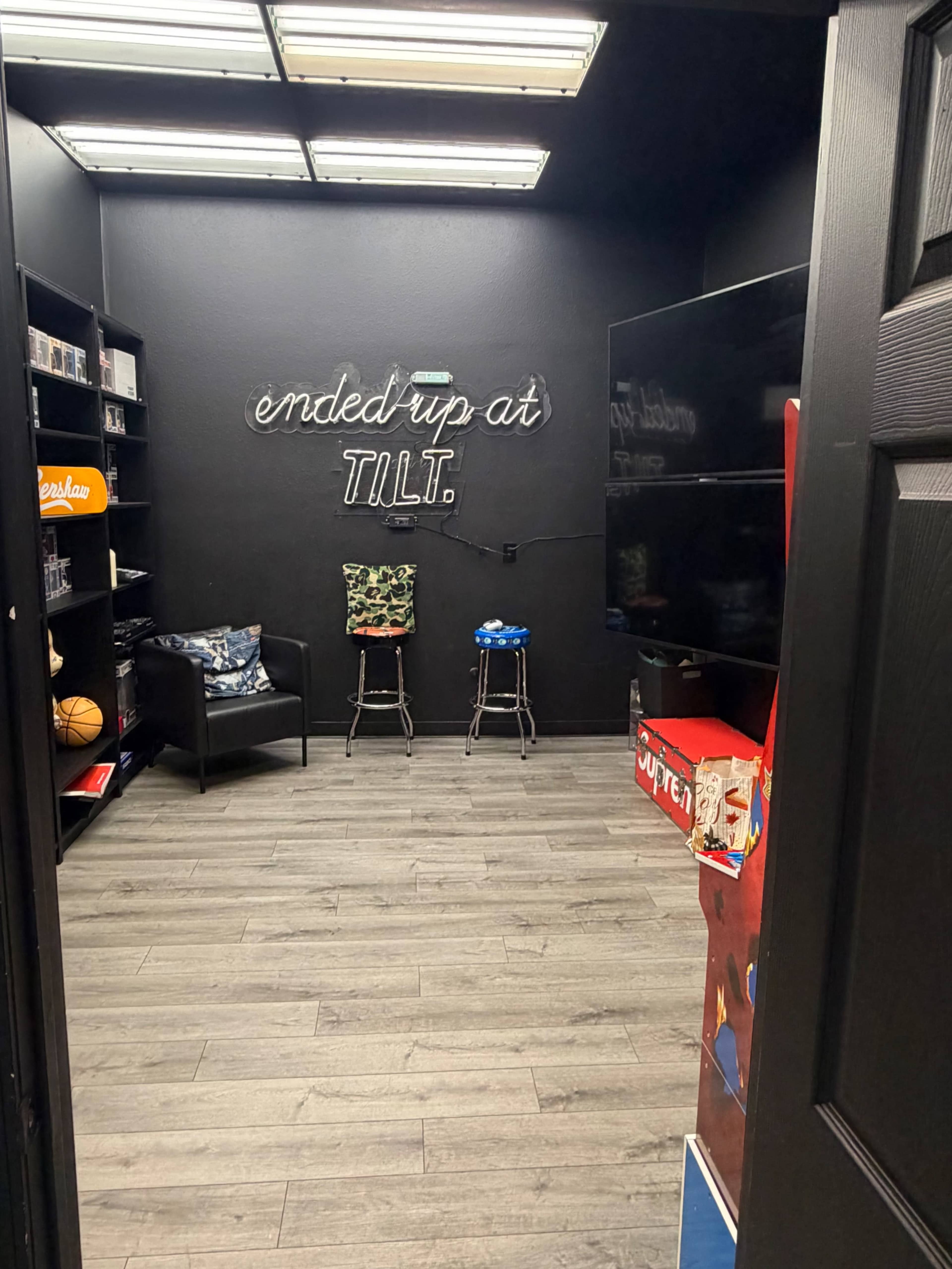 The image shows a small, dark room with shelves filled with items, two barstools, and a neon sign on the wall that says "ended up at TILT."