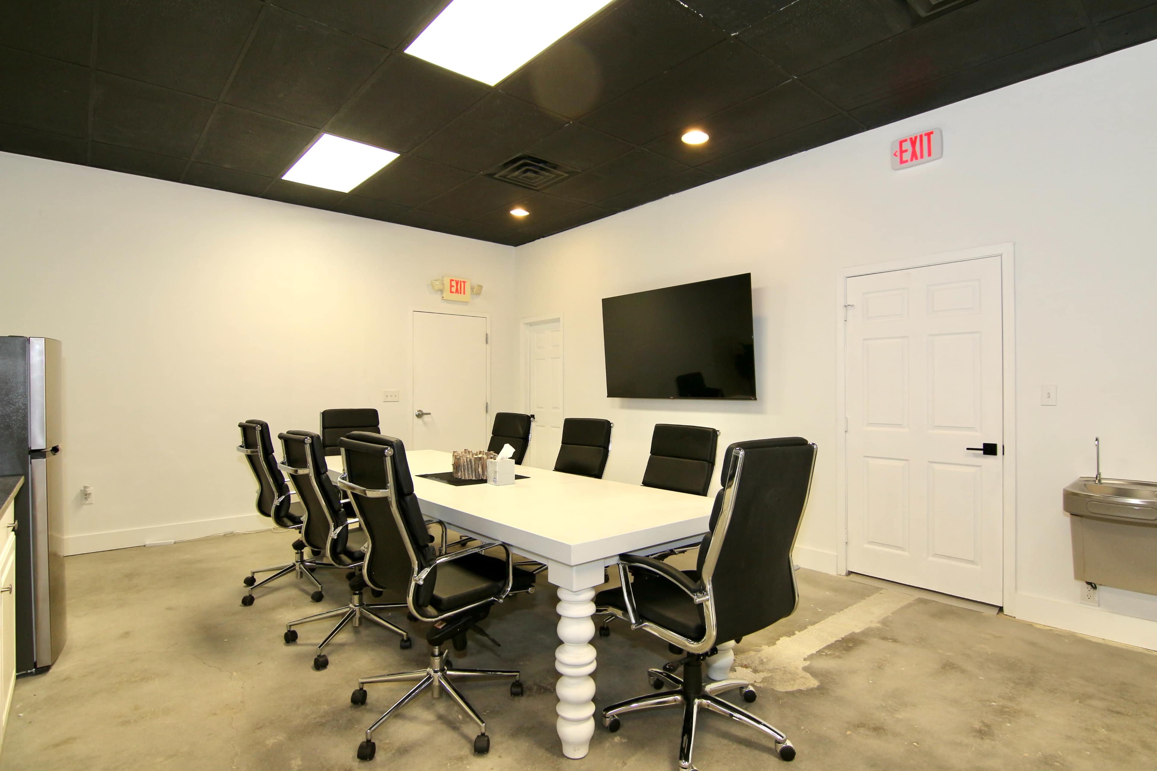 A conference room with a large table surrounded by black chairs, a wall-mounted television, and a door leading to another area.