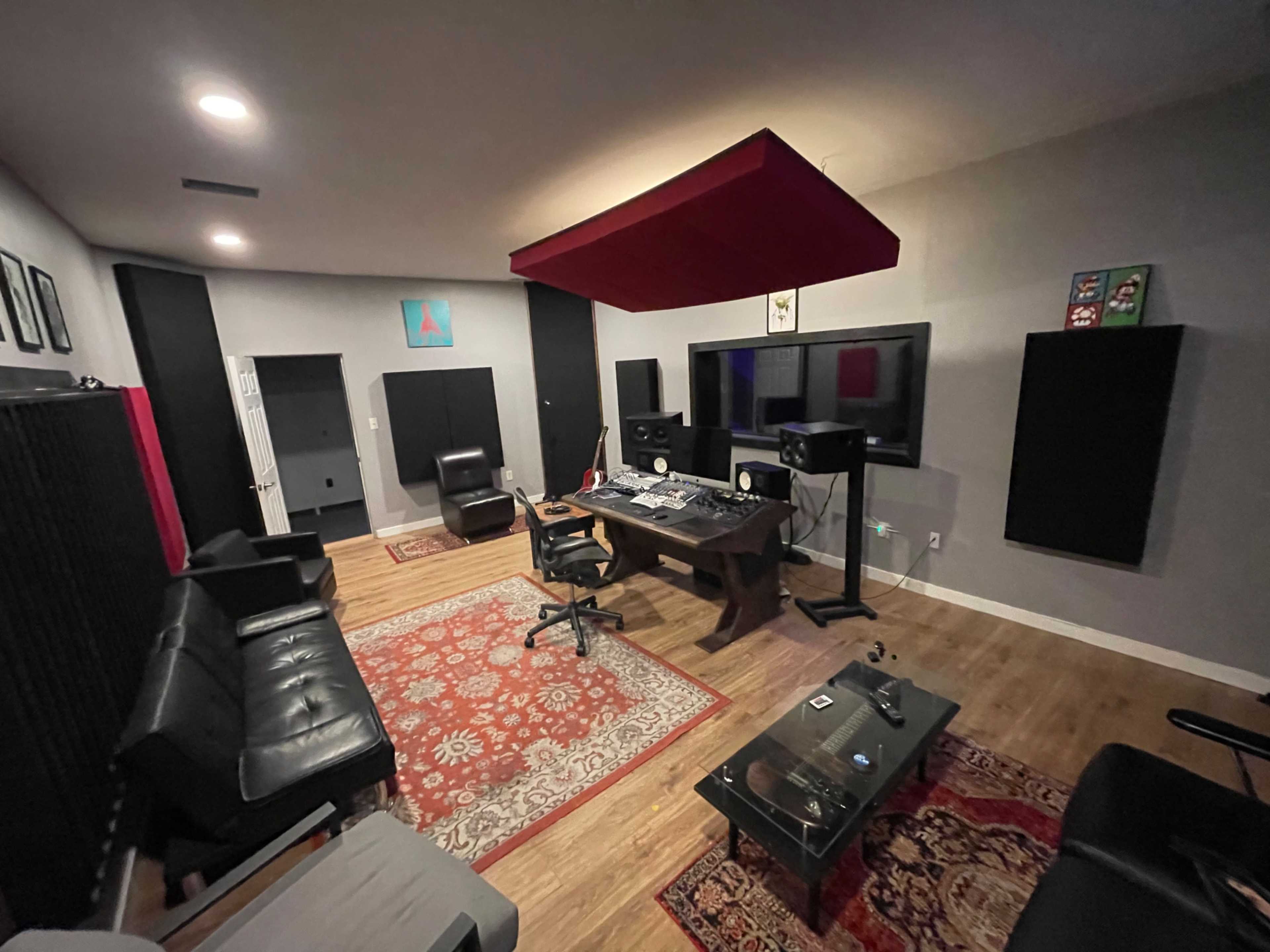The image shows a music studio with a control desk, chairs, soundproofing panels, and a large screen in a room with a brown wooden floor.