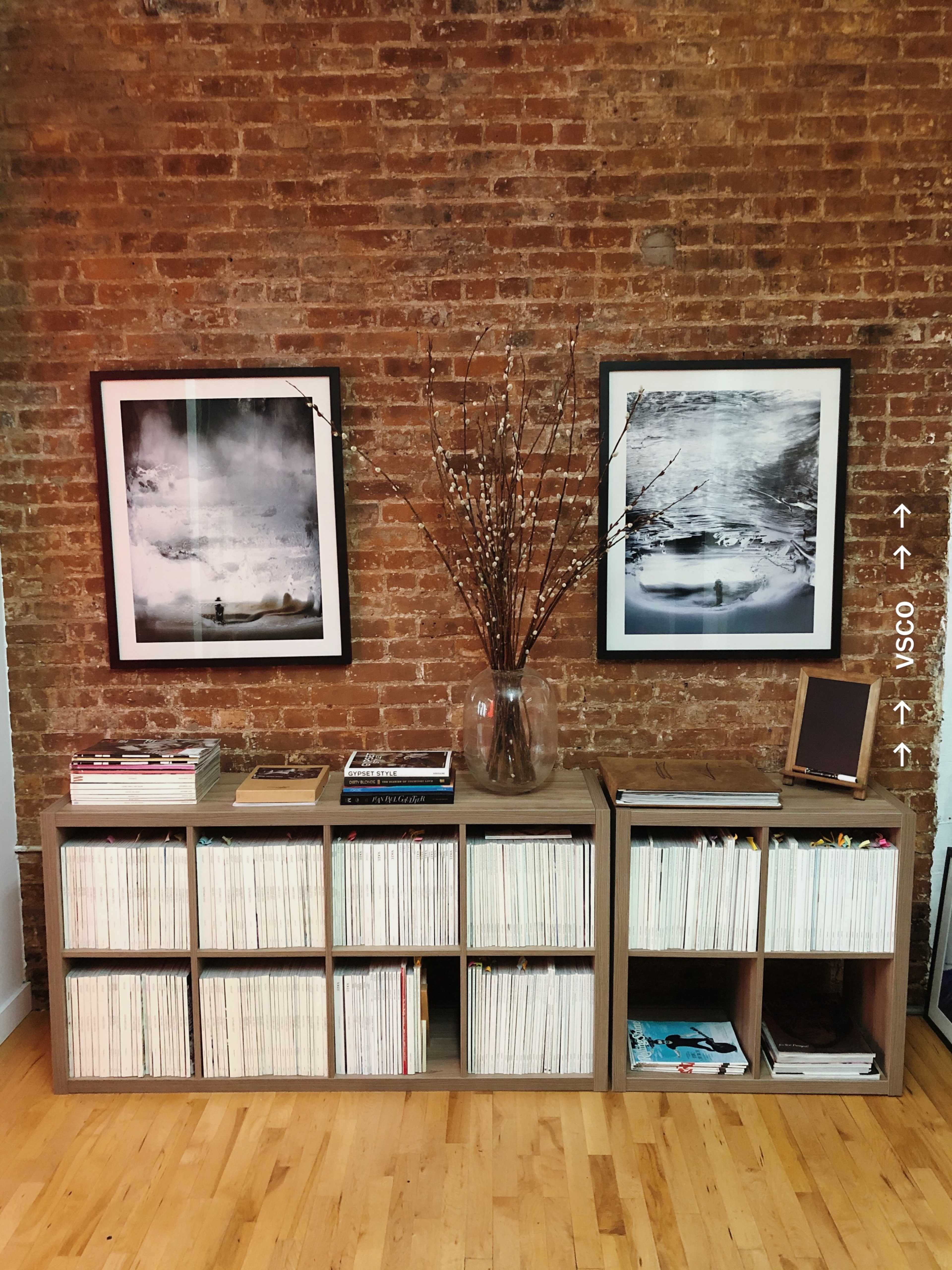 A wooden bookshelf filled with vinyl records and magazines is placed against a brick wall, with two framed black-and-white photographs and a vase of branches above it.