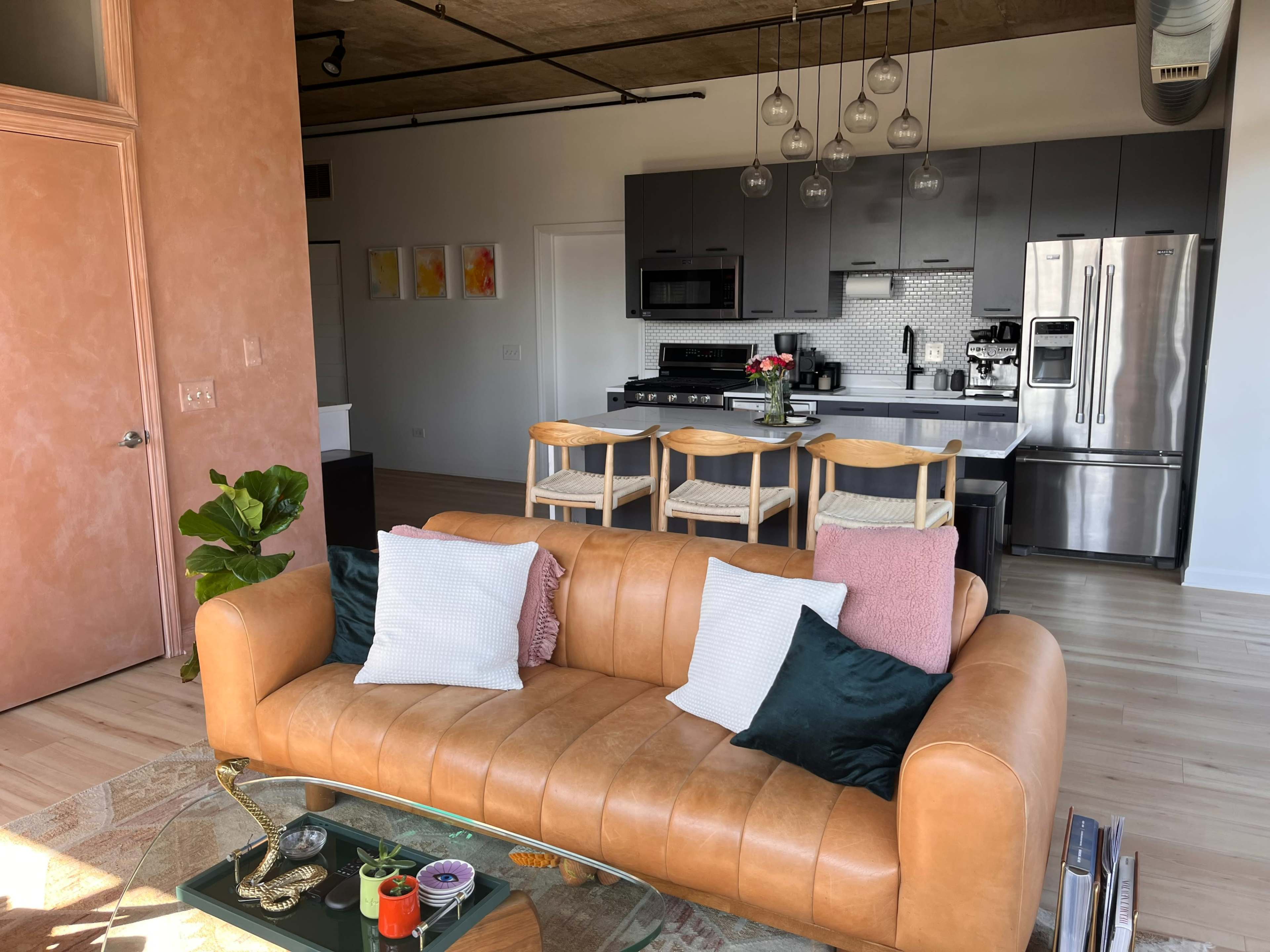 A modern living room with a tan leather couch, a glass coffee table, and a kitchen area featuring dark cabinets and a breakfast bar with wooden stools.