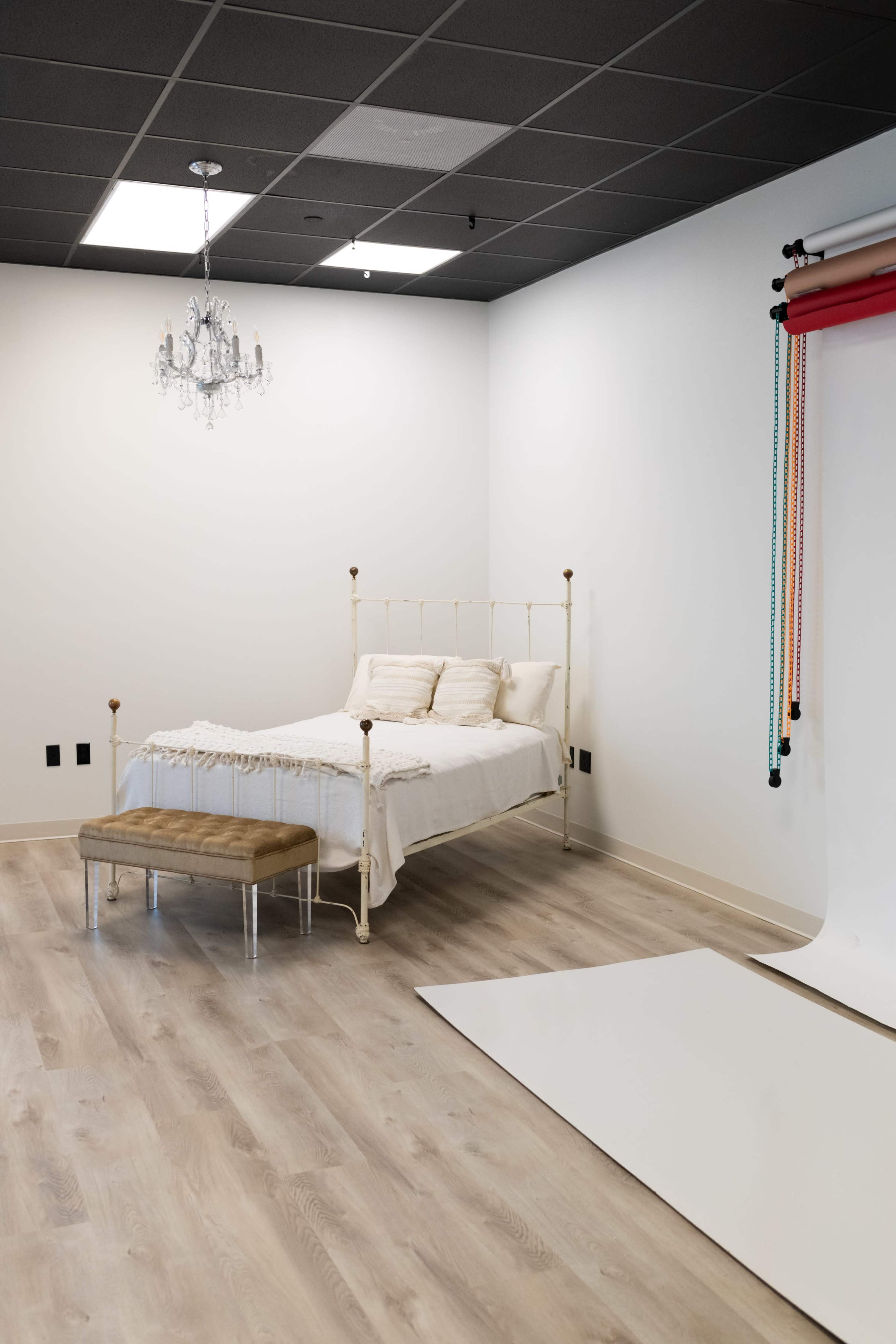 The image shows a minimalistic bedroom with a metal bed frame, a bench at the foot of the bed, and a backdrop setup, featuring a chandelier and colored rolls on the wall.