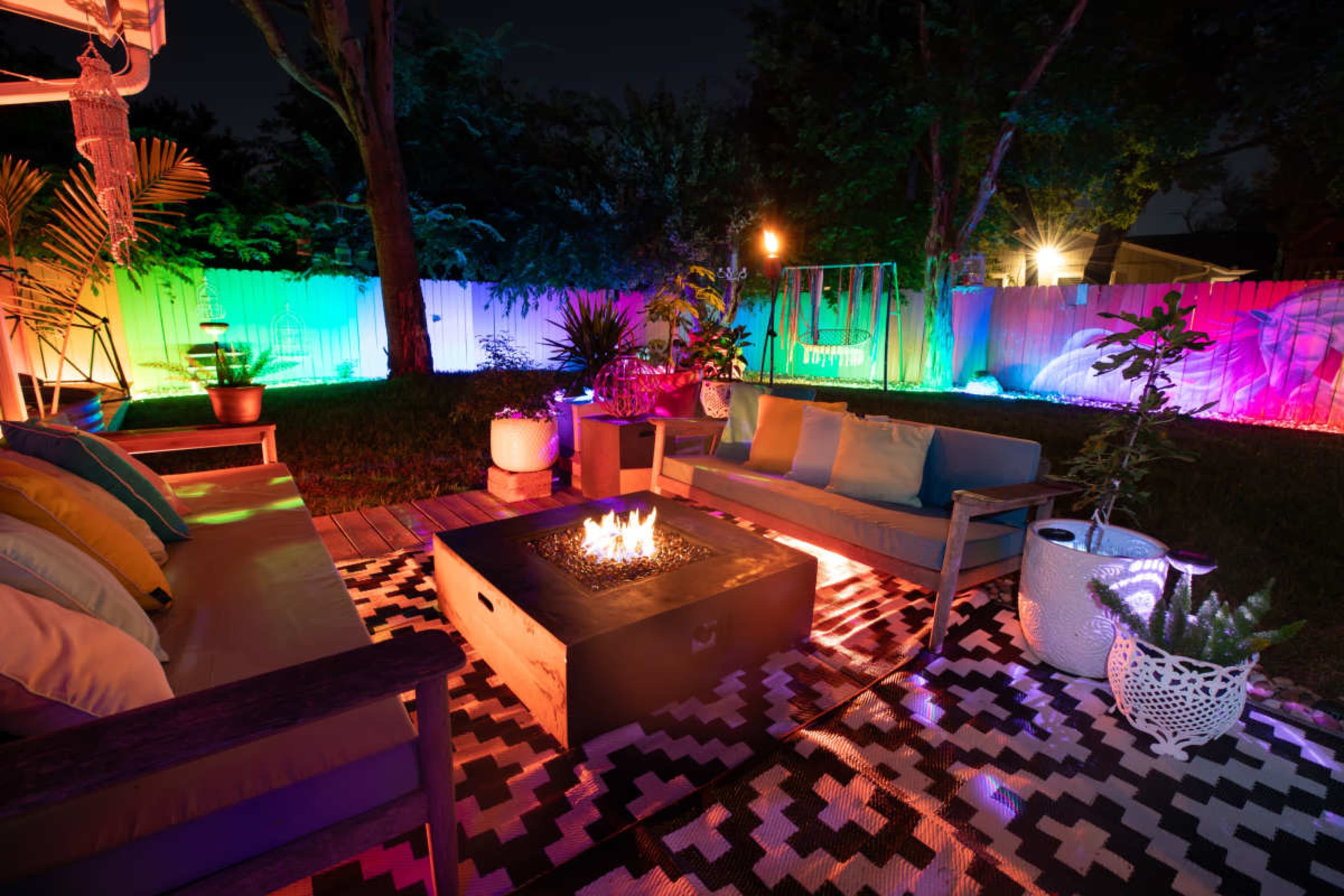 A modern outdoor seating area features a fire pit surrounded by cushions and colorful lighting against a backdrop of trees and a white fence.