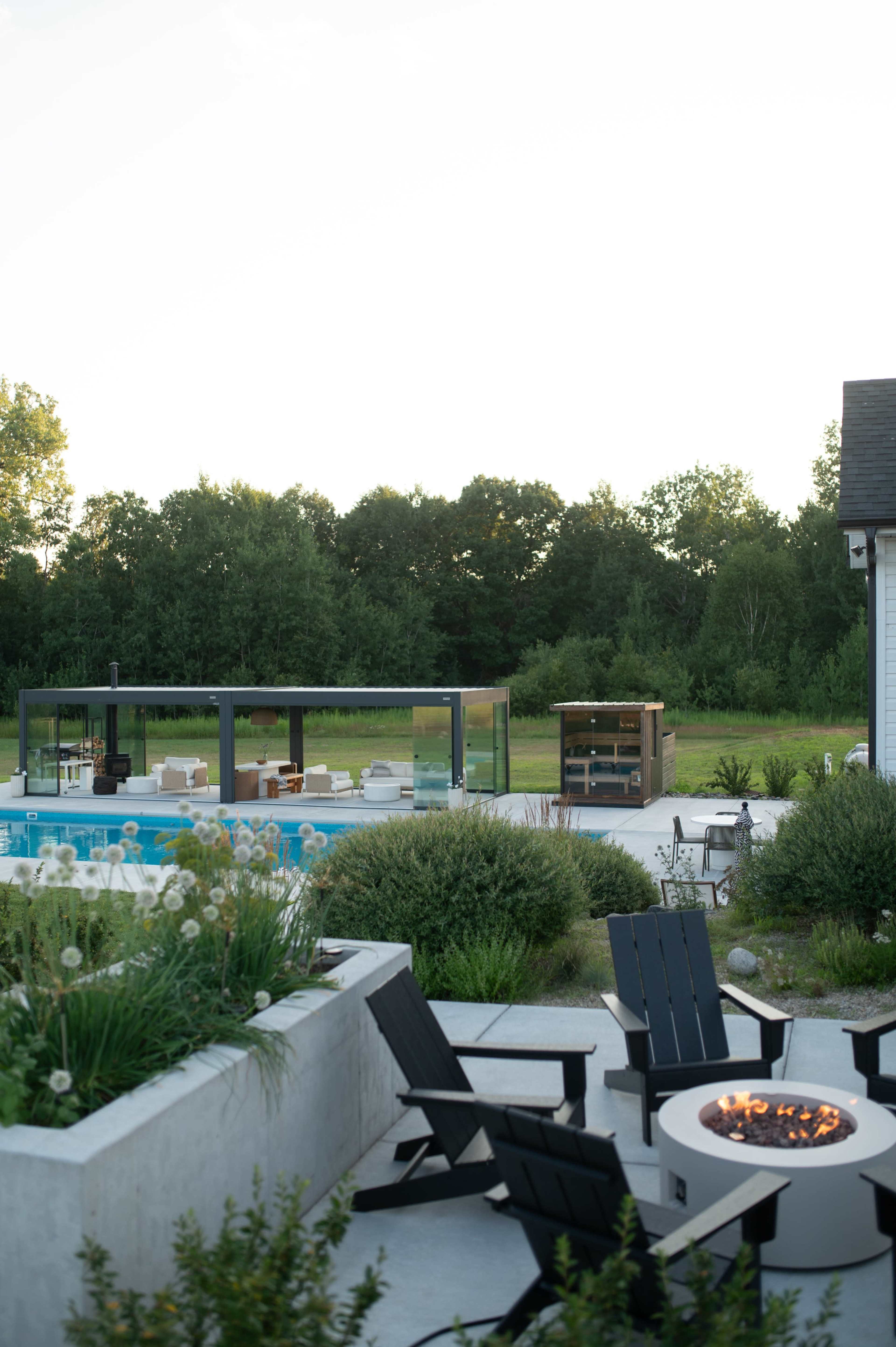 A modern outdoor space features a pool, a glass-walled lounge area, and a patio with fire pit chairs surrounded by greenery.