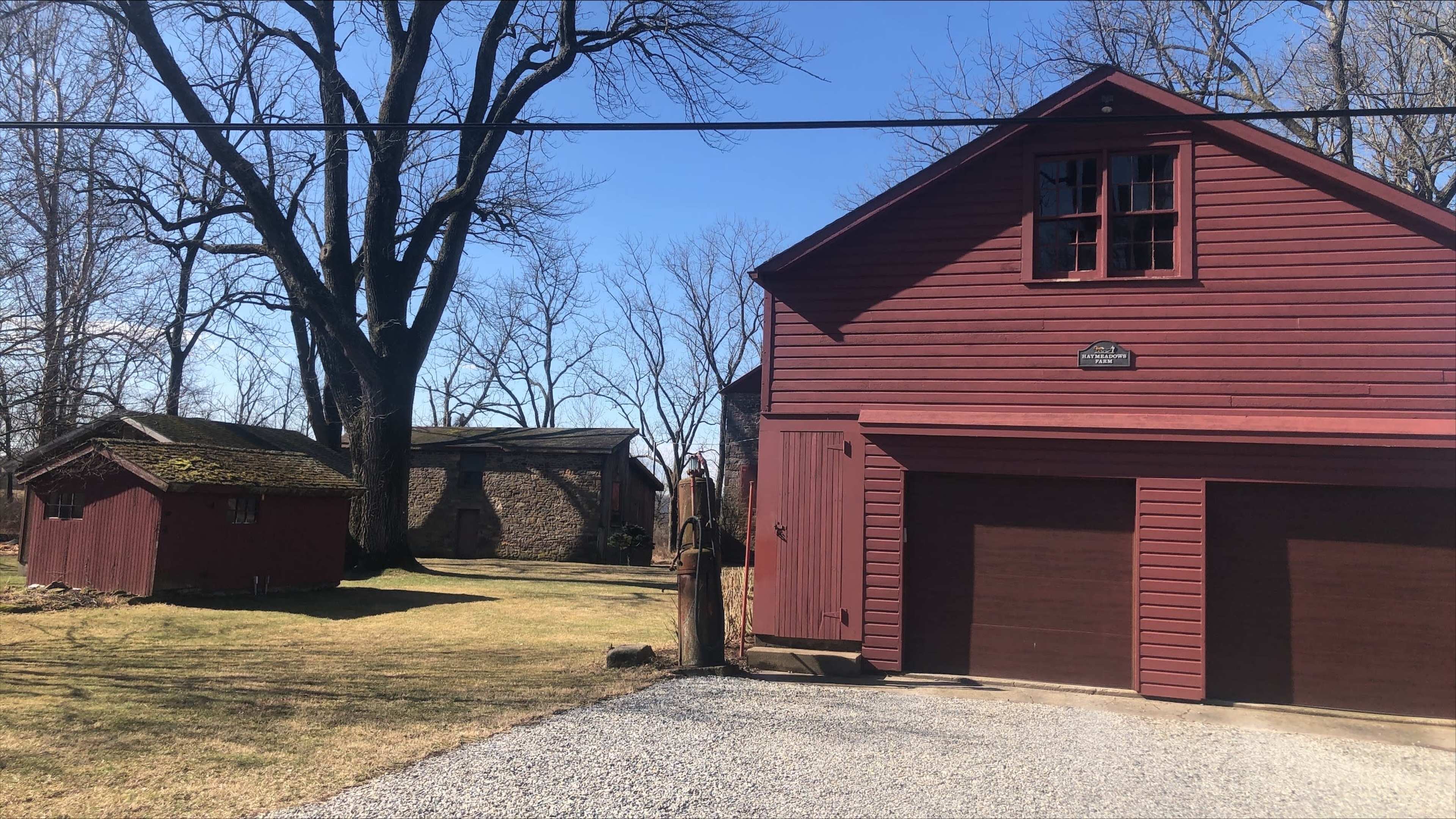 The image shows a red barn-style garage next to a smaller shed and a stone building, all set in a grassy area with bare trees under a clear blue sky.