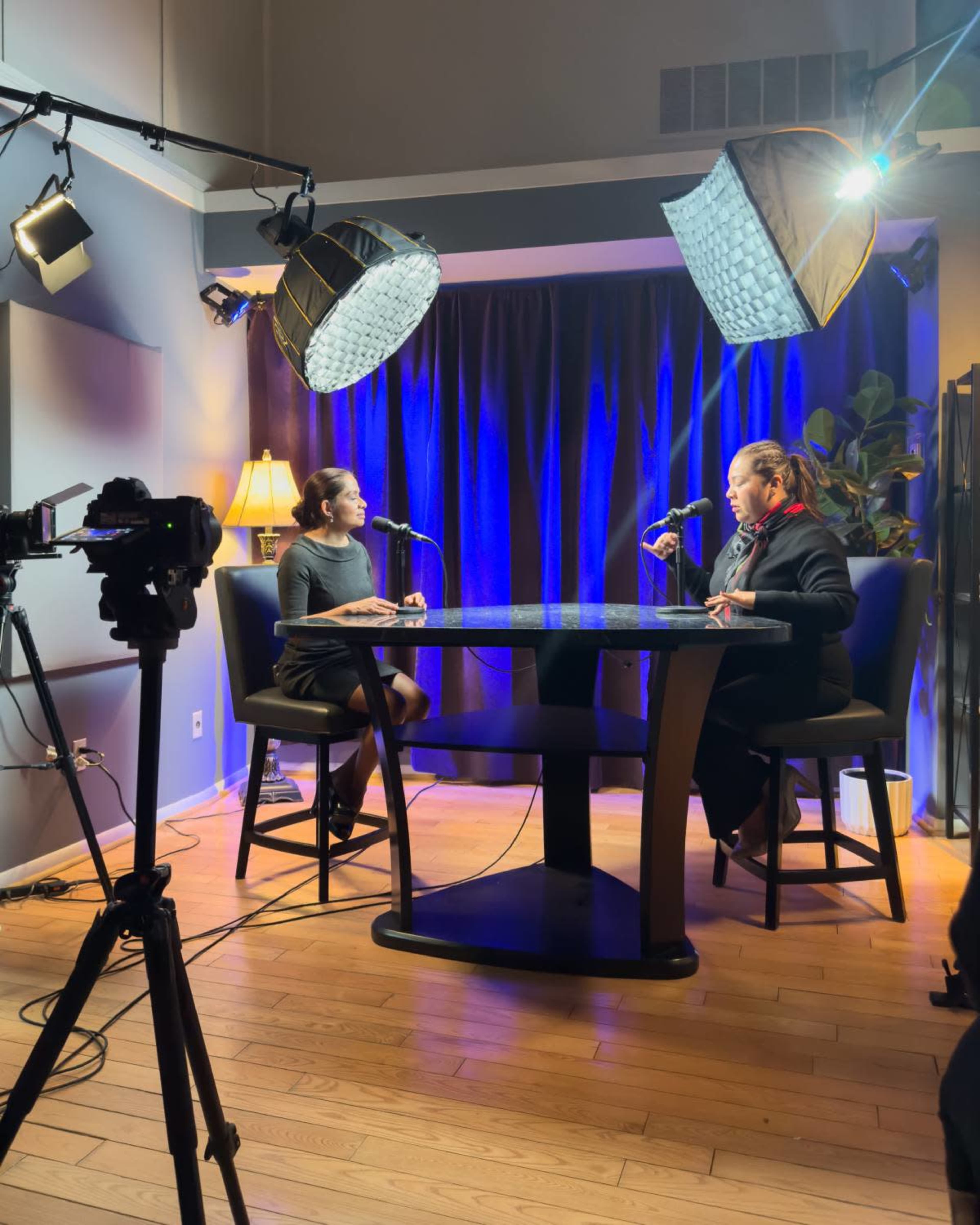 Two people are seated at a round table in a well-lit studio with blue backdrops, each holding a microphone.