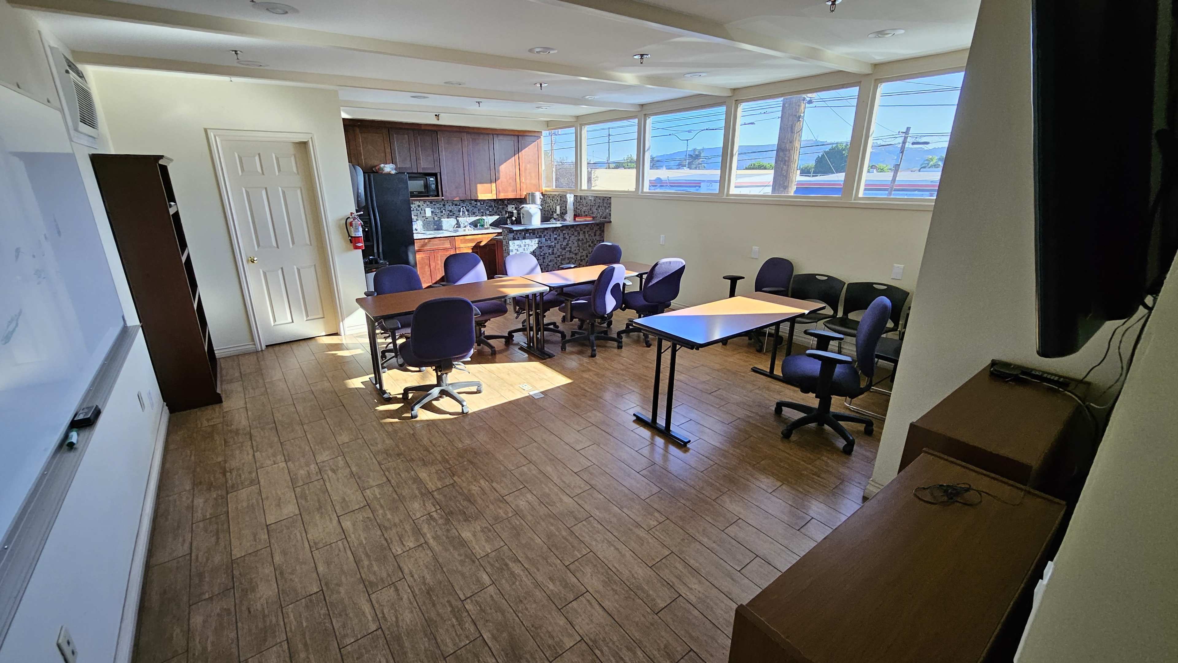 A classroom or meeting room with several tables and chairs arranged around a central area, along with a kitchenette in the background.
