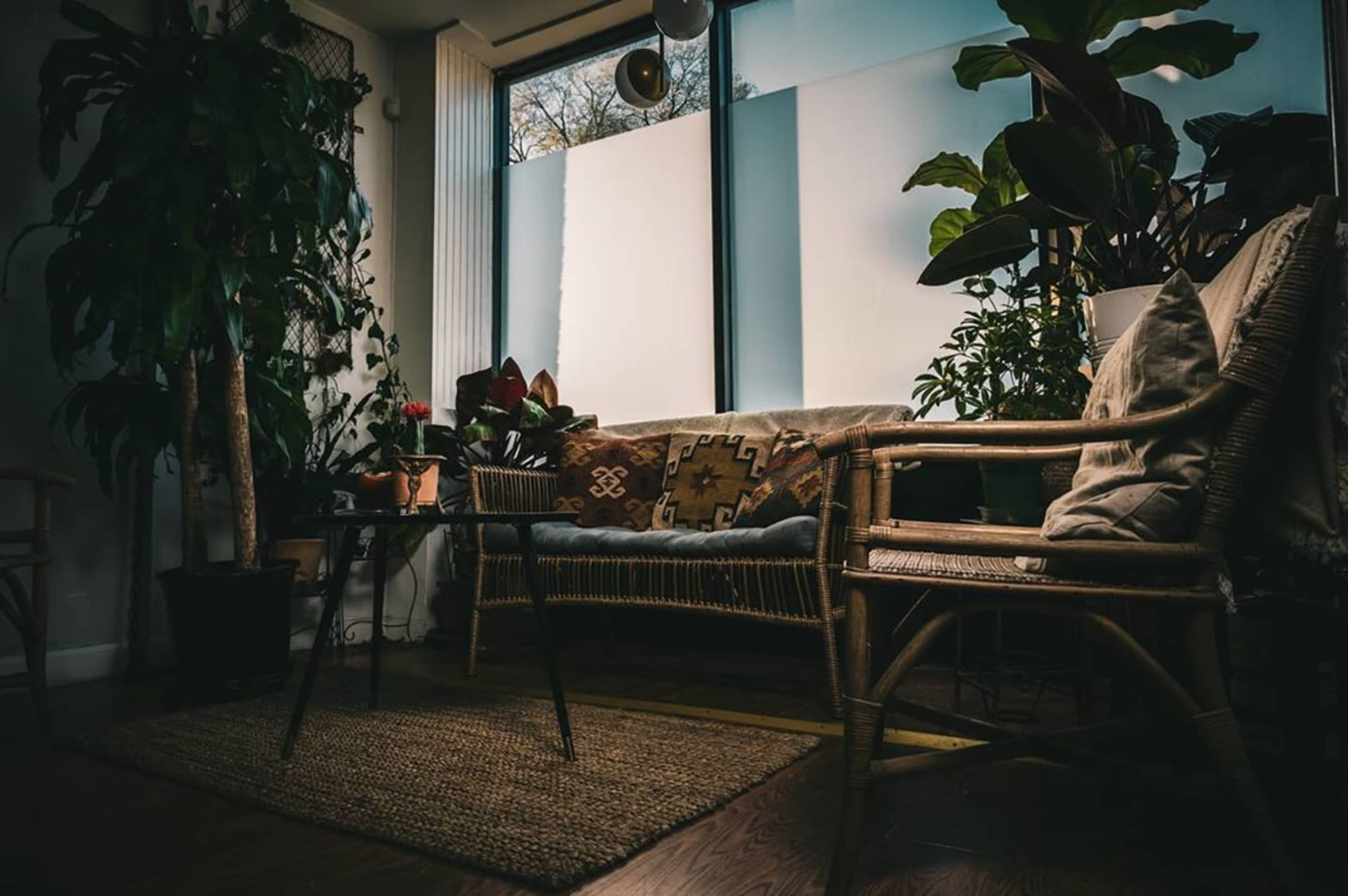 A cozy indoor seating area features a rattan chair, a small table, and various plants, with large windows providing natural light.