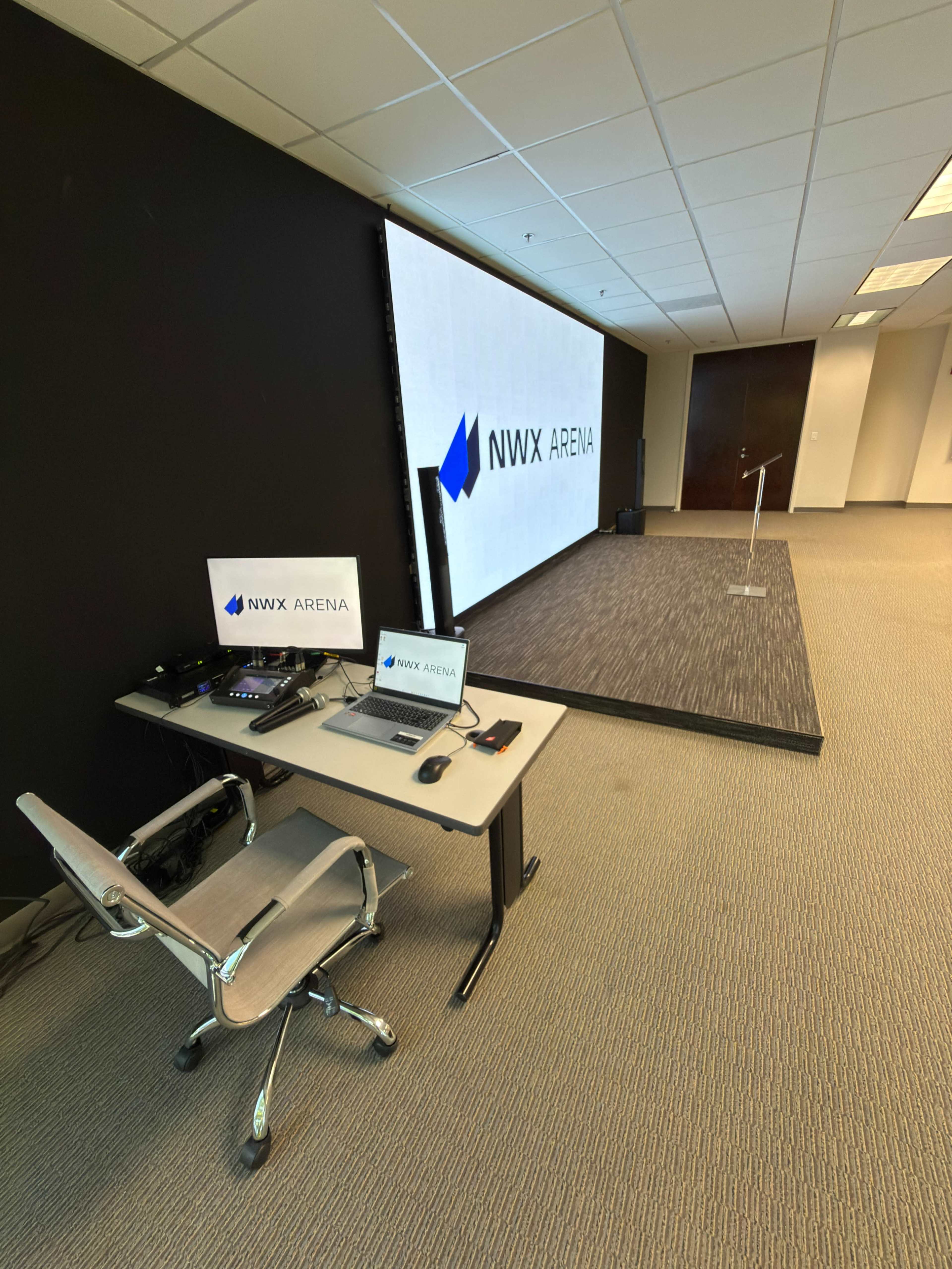A modern office space features a presentation area with a large screen displaying "NVX ARENA" and a desk equipped with a laptop and video equipment.