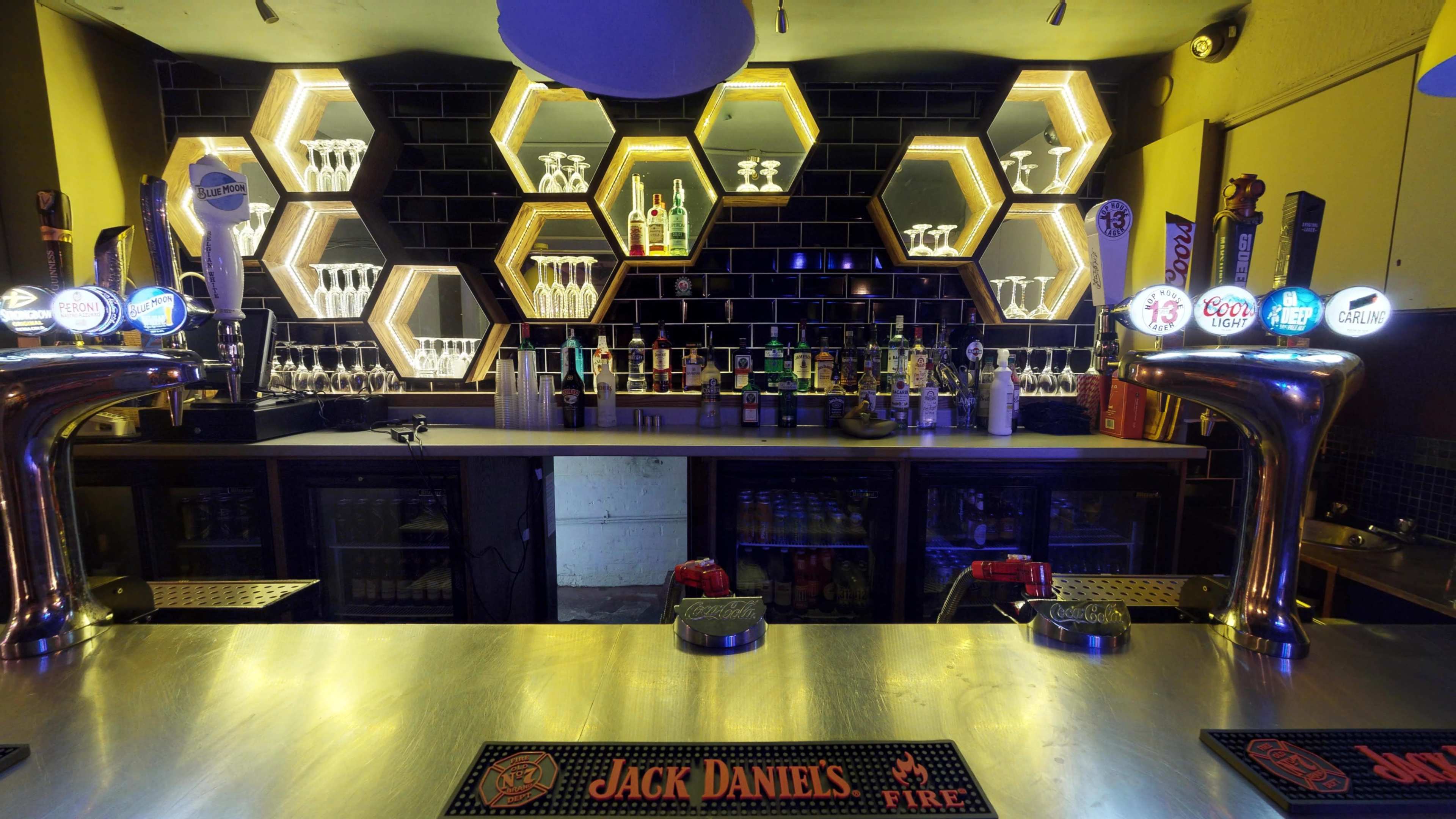 A well-lit bar features a honeycomb-shaped shelving unit displaying various bottles and glassware against a dark backdrop.